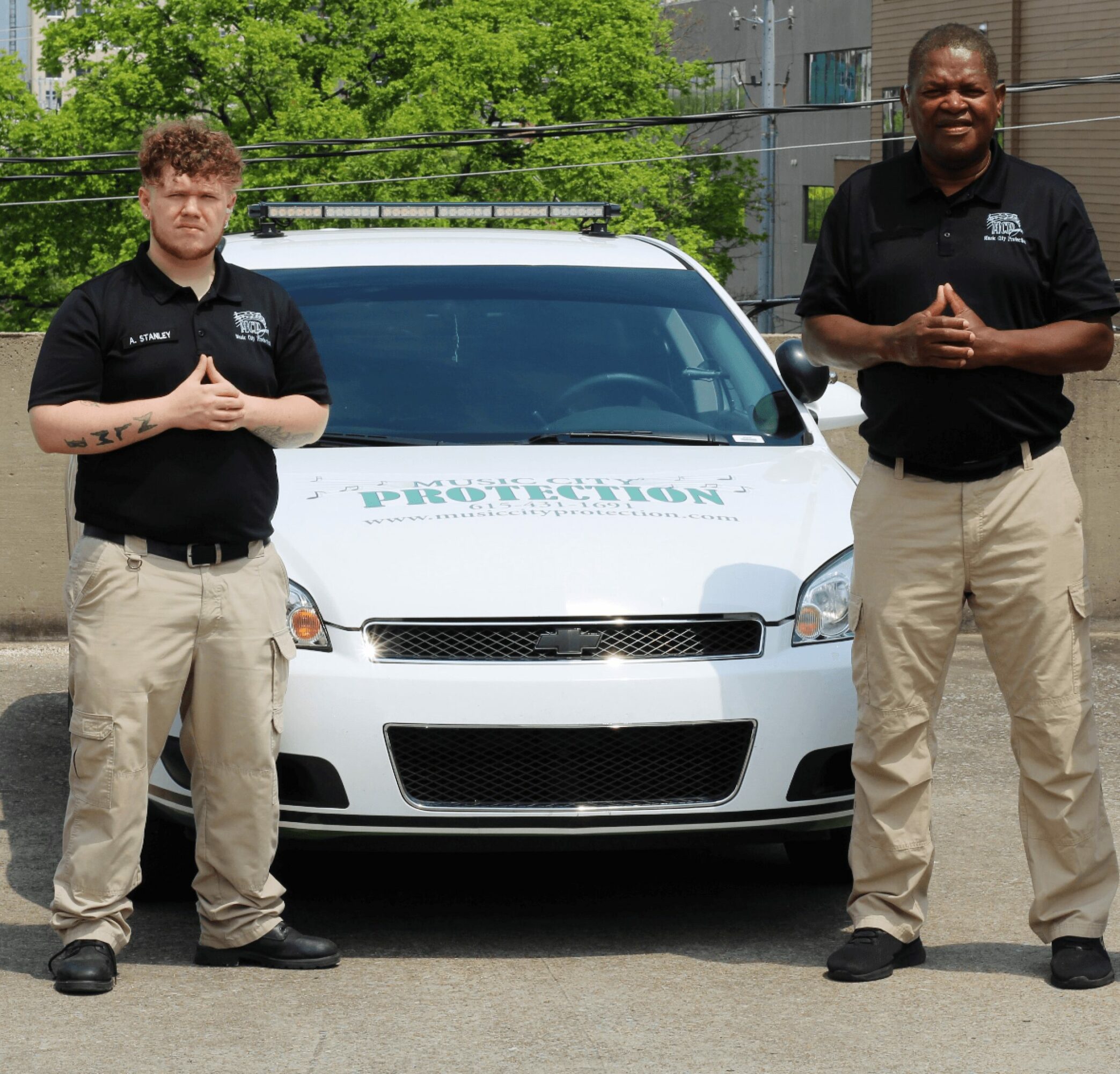 Two men stand in front of a white vehicle with a logo, outdoors on a sunny day, both with hands clasped.