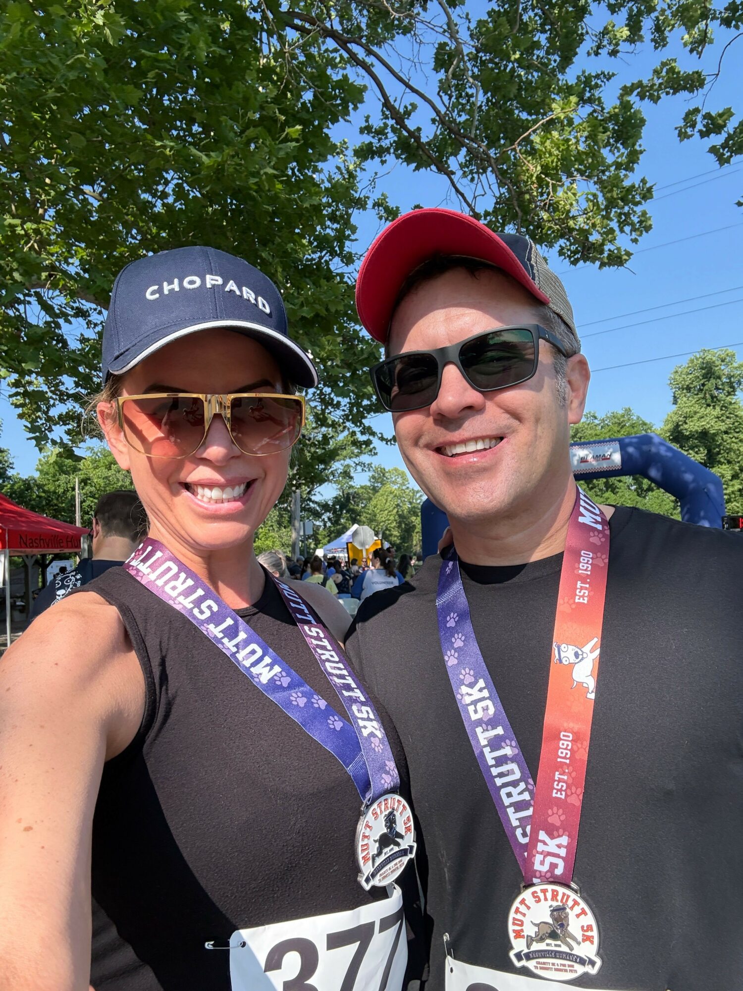 Two people smiling outdoors, wearing sunglasses and race bibs, with trees and blue sky in background.