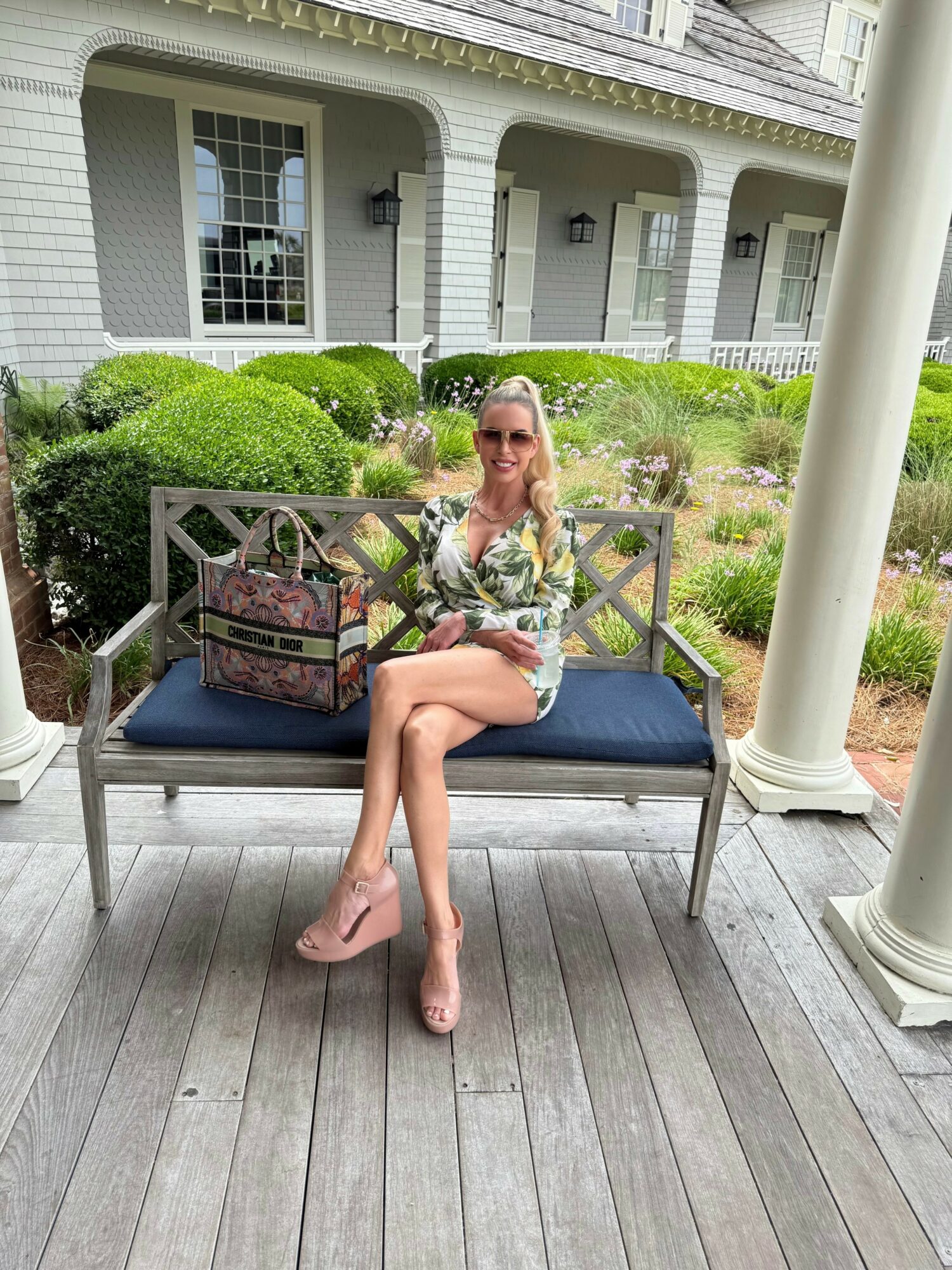 Woman sitting on a bench outdoors, holding a drink, with a house and garden in the background.