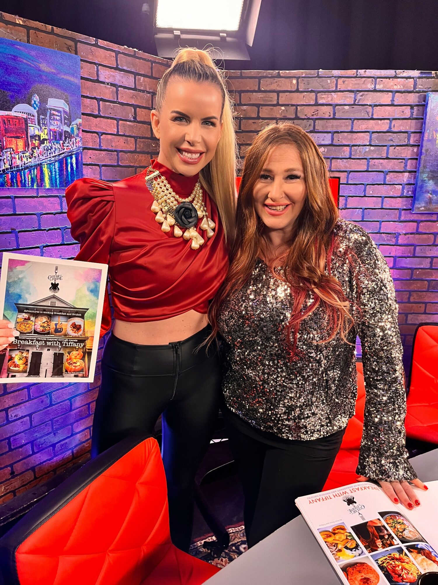 Two women smiling indoors, one holding a colorful poster, brick wall background, studio lighting, seated at a table.