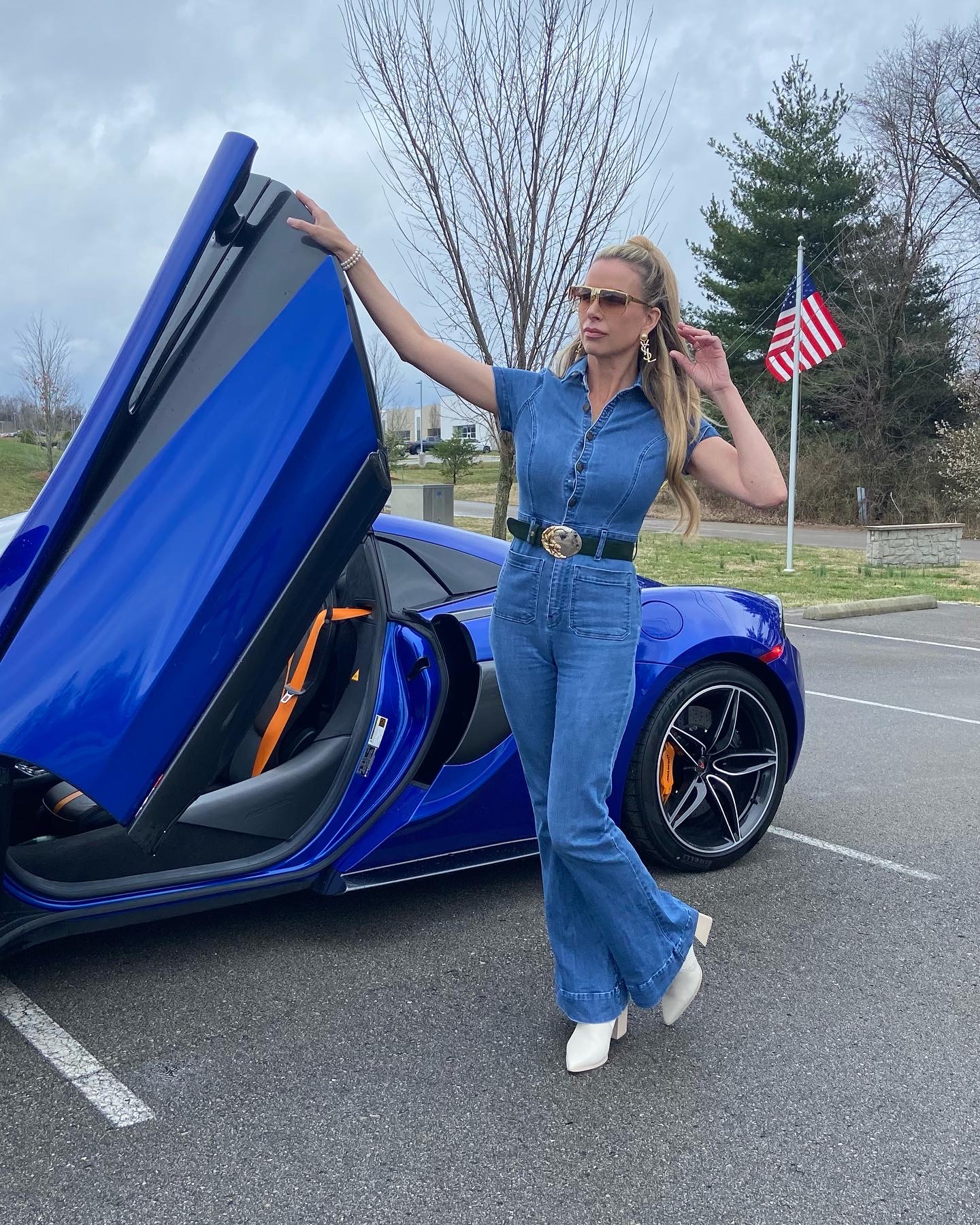Woman in blue jeans and top standing next to a blue sports car with open door, in a parking lot with American flag and trees.
