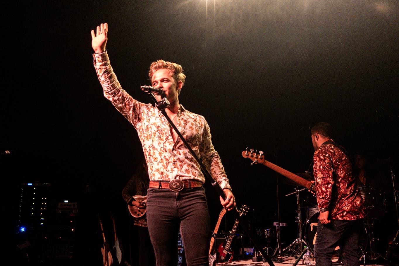 Person on stage waving with one hand, holding a microphone, with a band in the background, dark stage lighting.