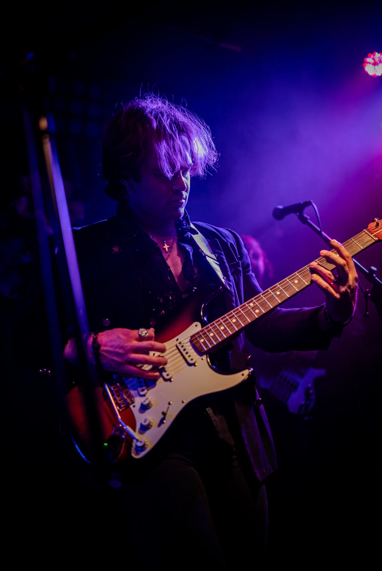Person playing an electric guitar on stage with purple and blue lighting, hair covering face, microphone nearby.
