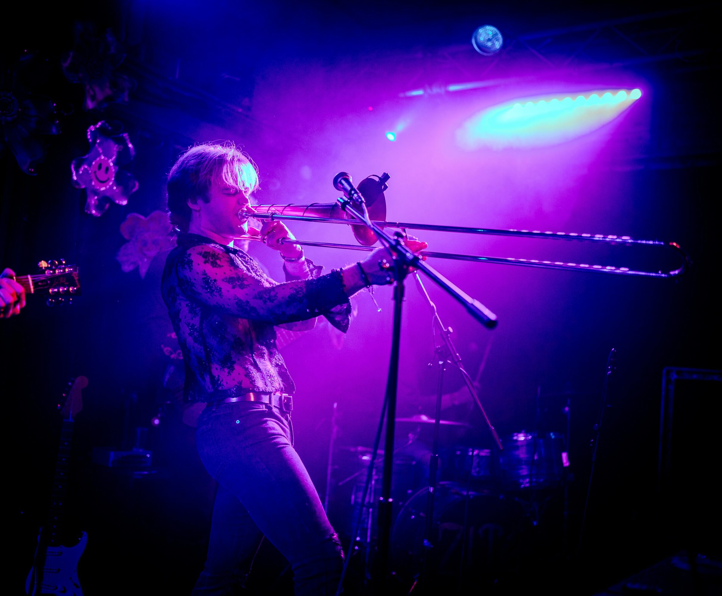 Person playing a wind instrument on stage with purple and blue lighting, microphone stand nearby.