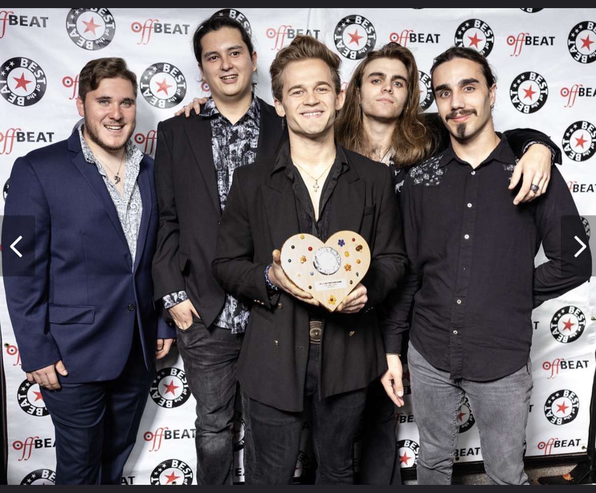 Group of five men standing together, smiling, with a backdrop featuring logos and text, one holding a heart-shaped award.