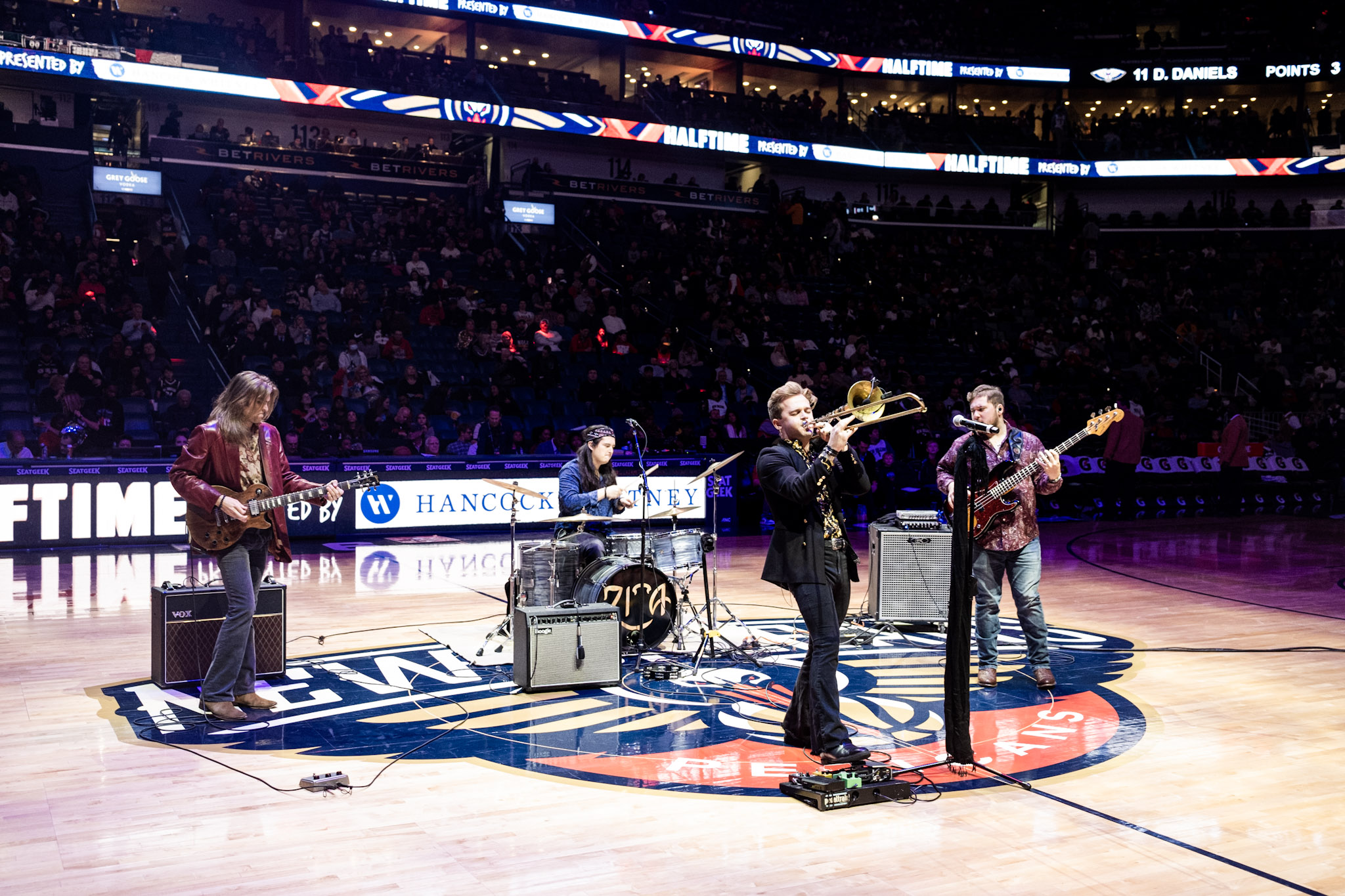 Band performs on basketball court with audience in background, stage setup includes musical instruments and equipment, arena lighting.