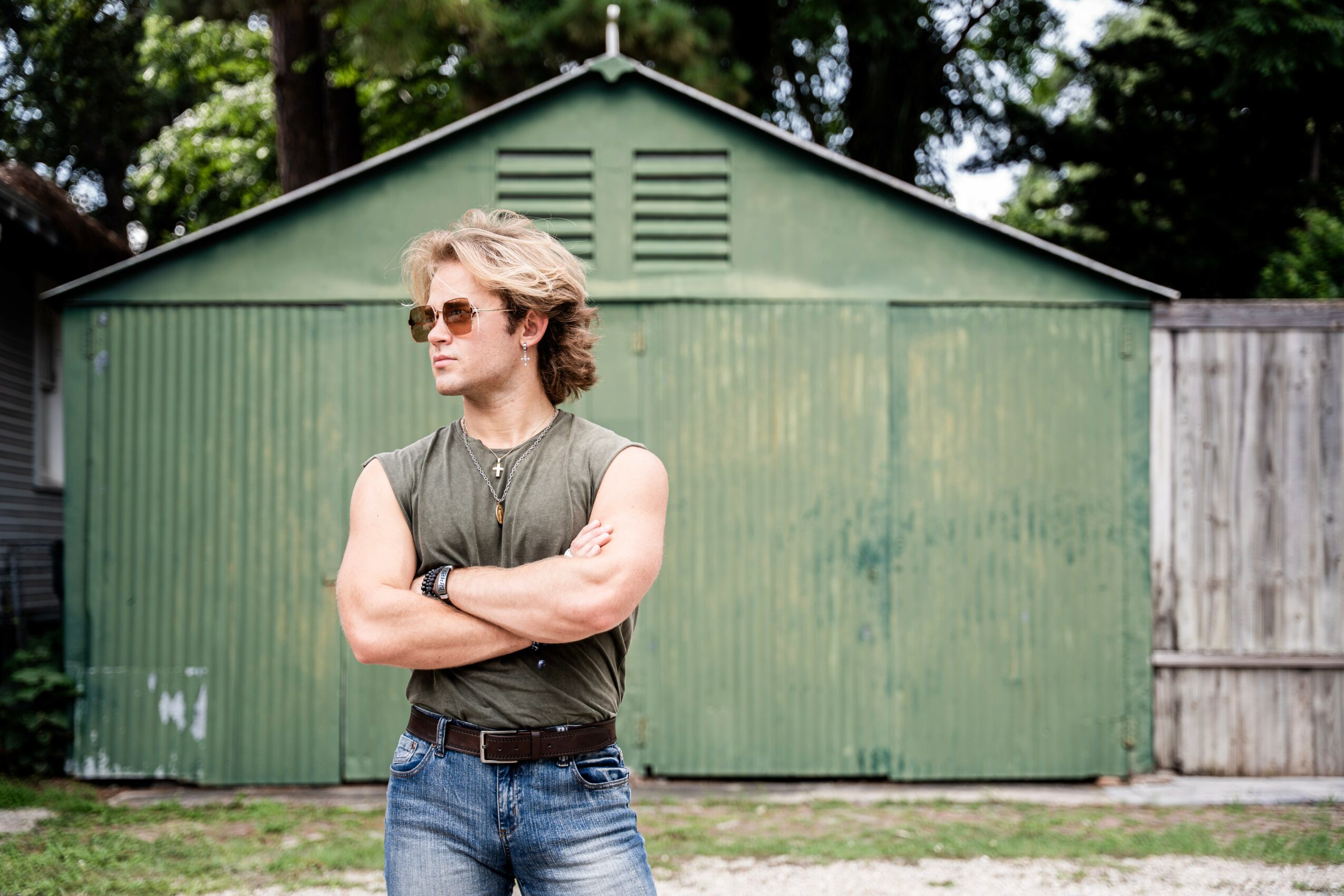 Person with sunglasses and curly hair standing with arms crossed in front of a green shed.