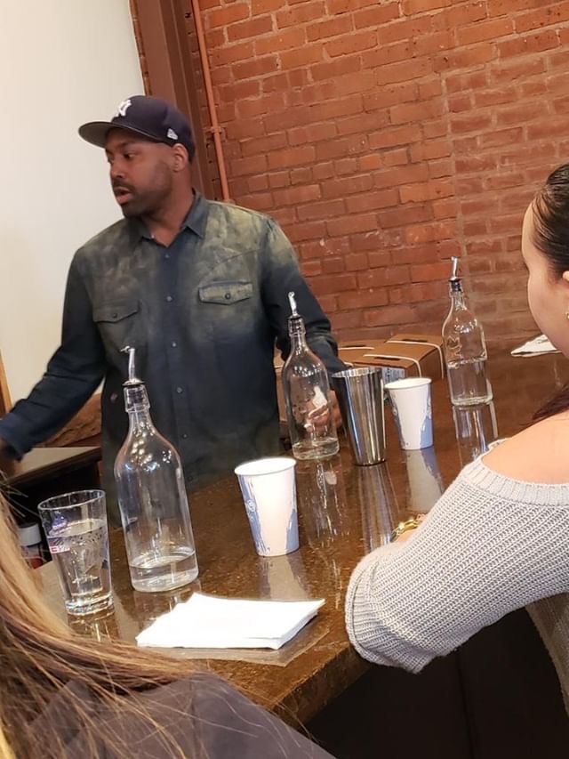Man in dark jacket and cap standing at a table with bottles and cups, talking to a group, brick wall background.