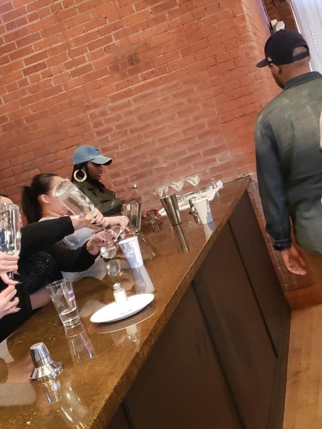 People sitting at a bar with glasses and bottles, brick wall background, one person wearing a cap, another with large earrings.