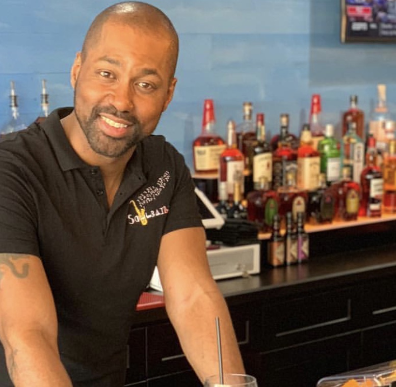 Man smiling behind bar with bottles of alcohol in background, wearing black shirt, in a bar setting.