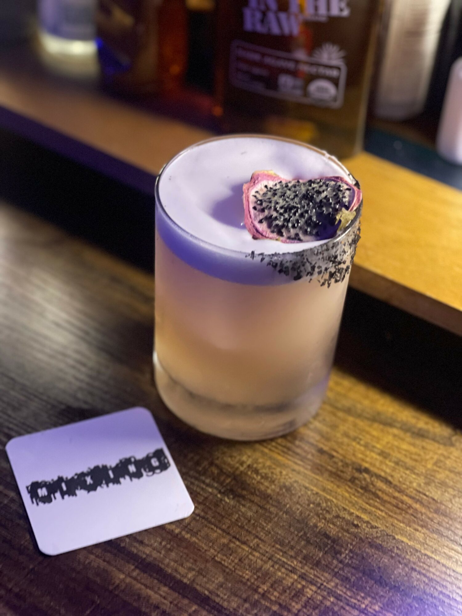 A layered cocktail in a glass with a purple top, garnished with a slice of fruit and black seeds, on a wooden table.