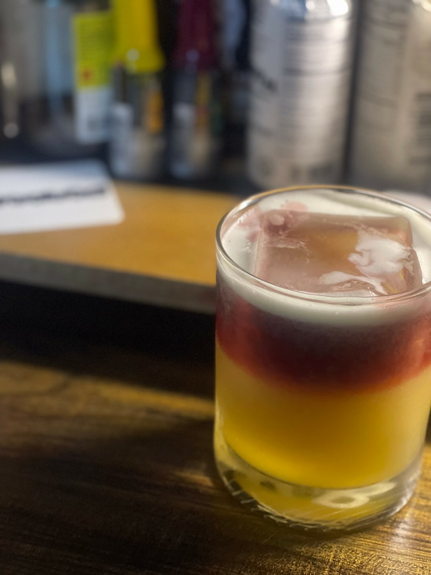 Layered drink with red, white, and yellow sections in a glass on a wooden surface, bar supplies in background.
