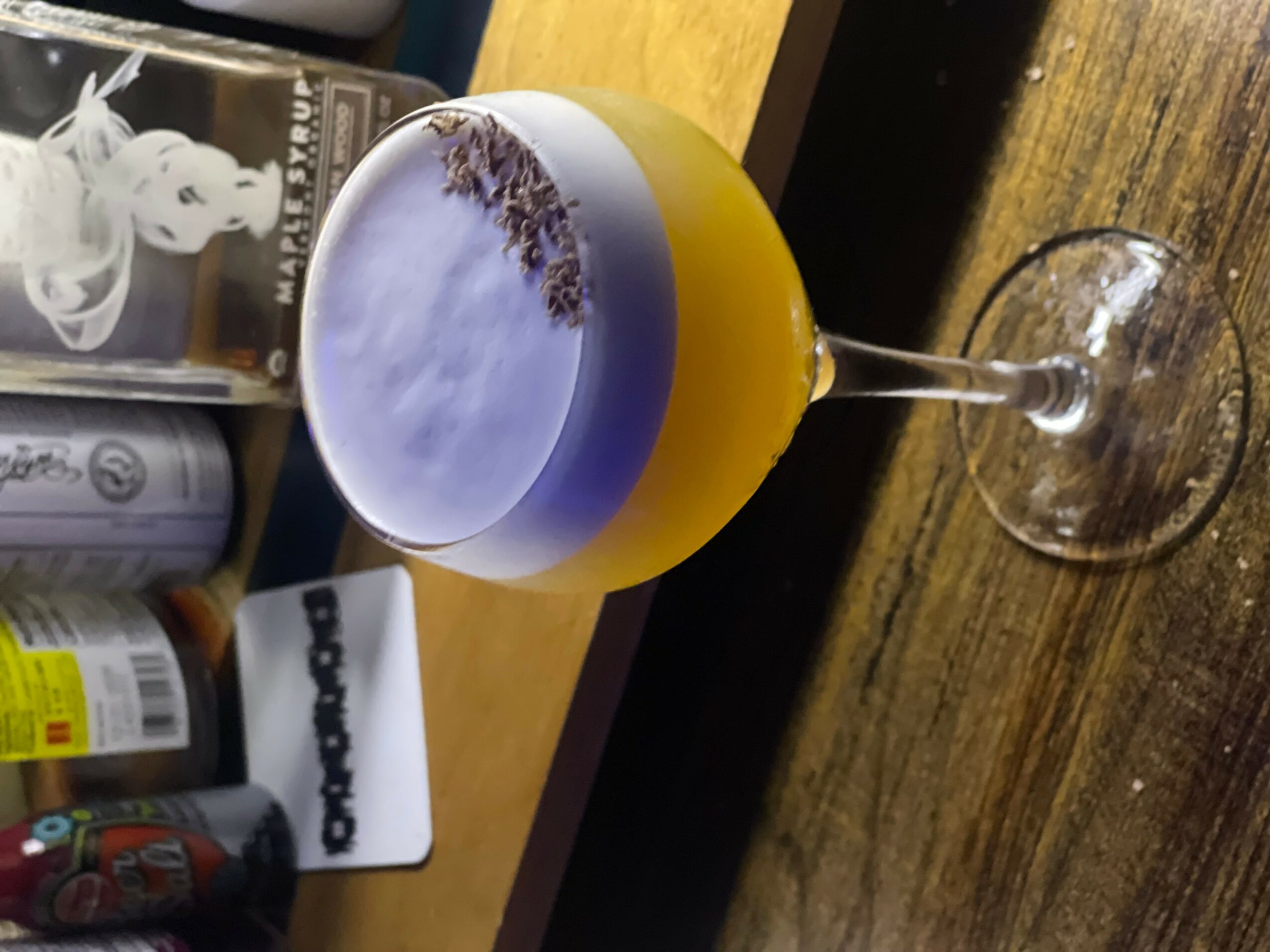 Cocktail glass with layered purple and yellow drink on wooden table, bottles and a book in background.