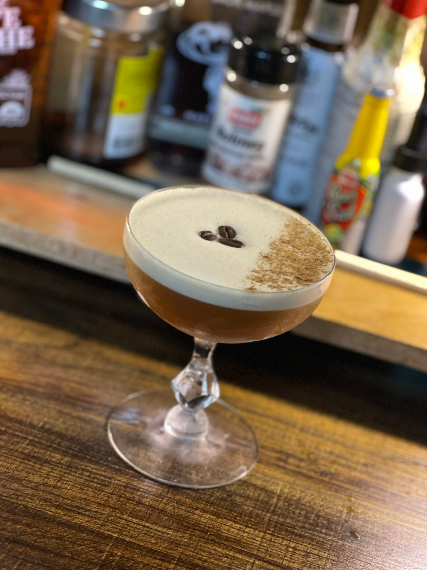 Coffee cocktail with foam and coffee beans on top, served in a glass on a wooden bar, background with bottles.