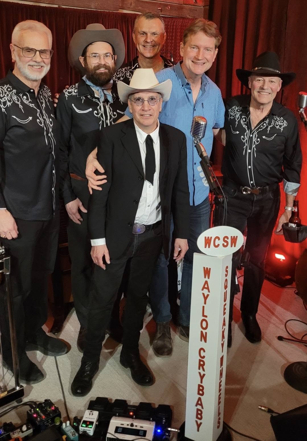 Six men in western attire pose on stage with a microphone and a sign reading 'WAYLONG CRYBABY'.
