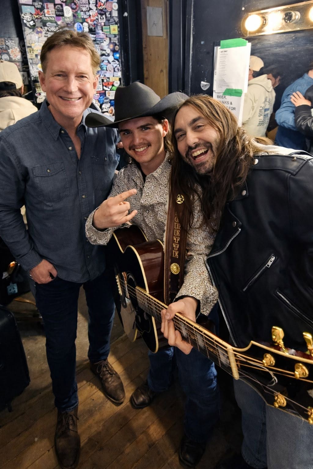 Three people smiling, one holding a guitar, in a cozy indoor setting with a crowd in the background.