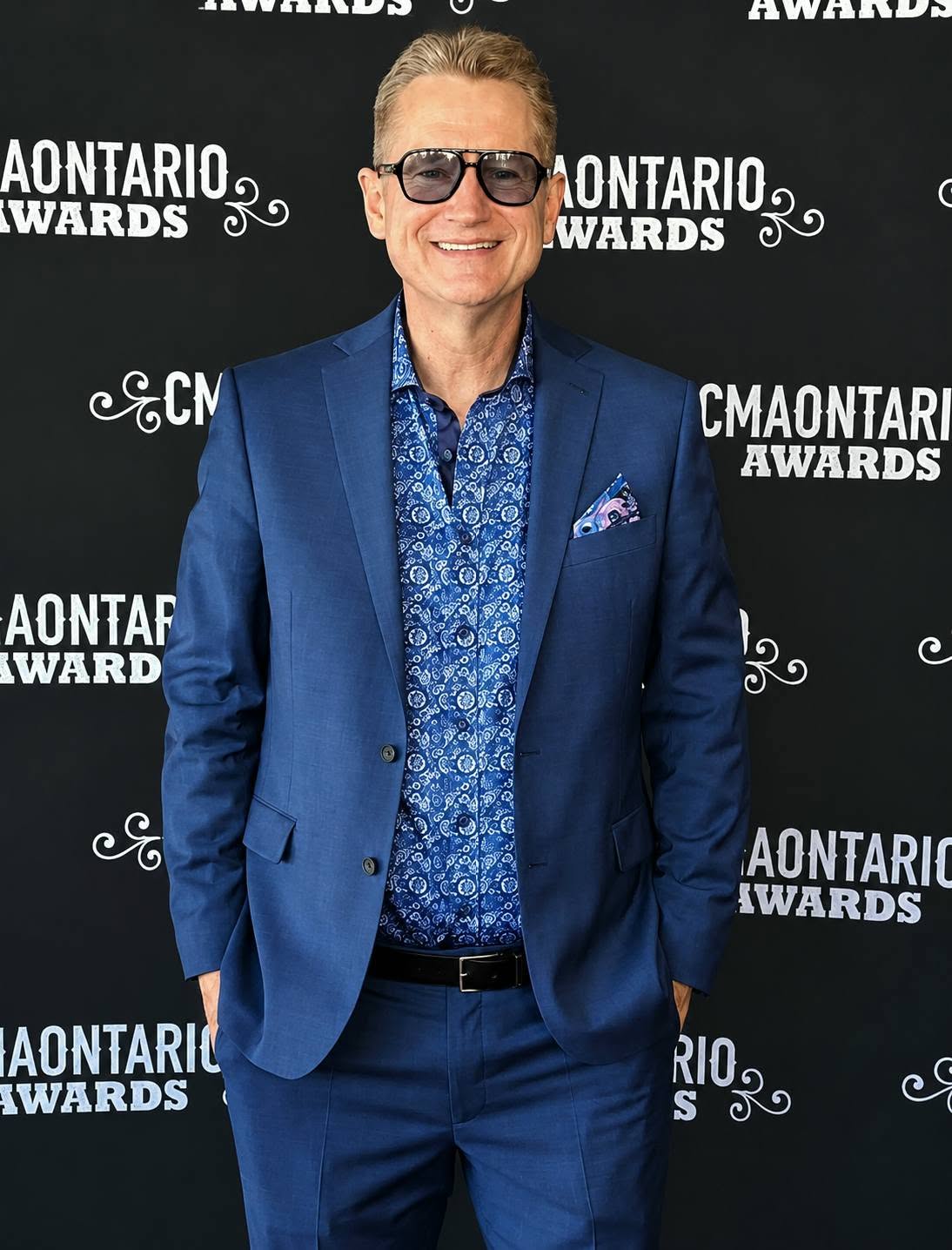 Man in a blue suit and sunglasses smiling at an event with a black background and white text.