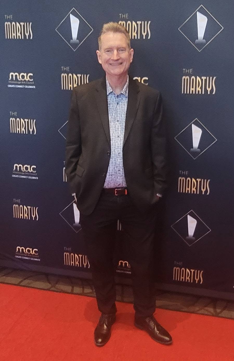 Man in a suit standing on a red carpet in front of a backdrop with event logos, smiling at the camera.