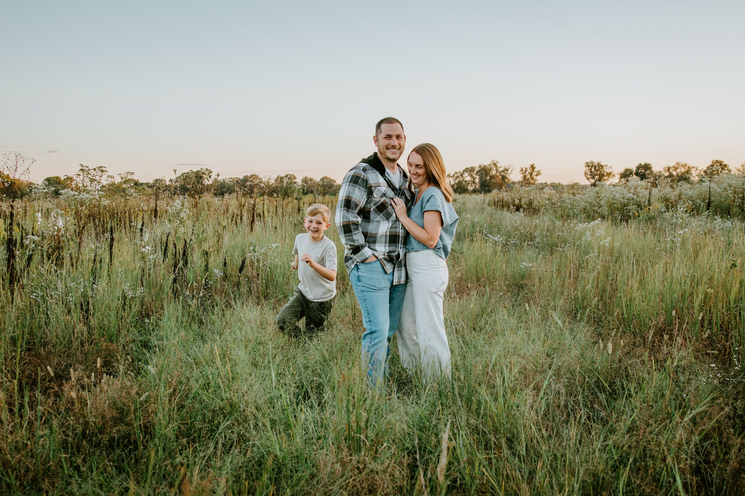 Family of three standing in a grassy field with a clear sky, smiling and embracing each other, child standing nearby.