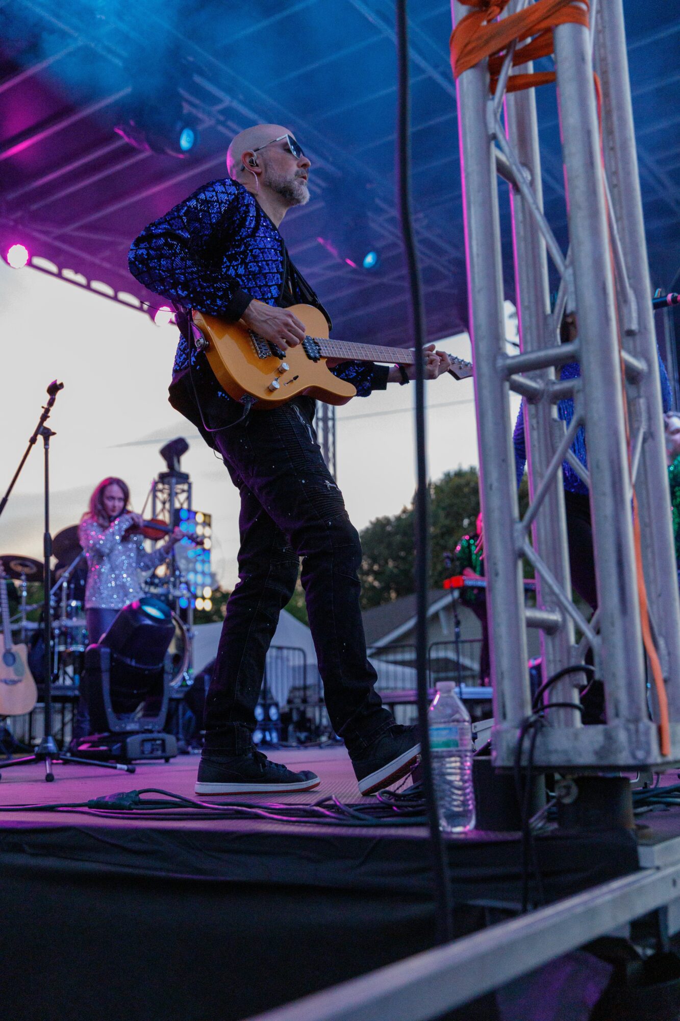 Musician playing guitar on stage during outdoor concert with band members and equipment visible.