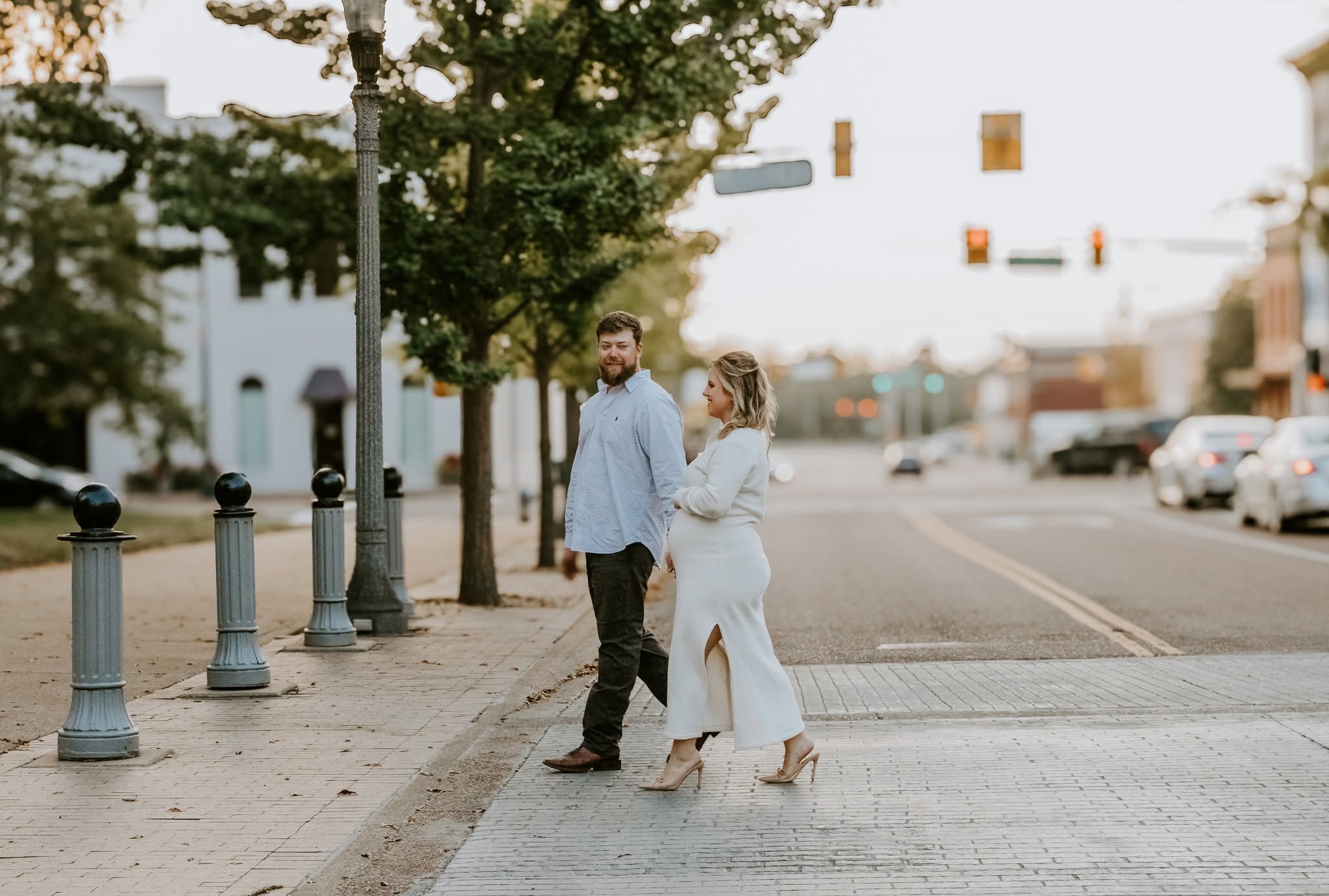 A man and woman stand on a city sidewalk, holding hands and looking at each other, with trees and buildings in the background.