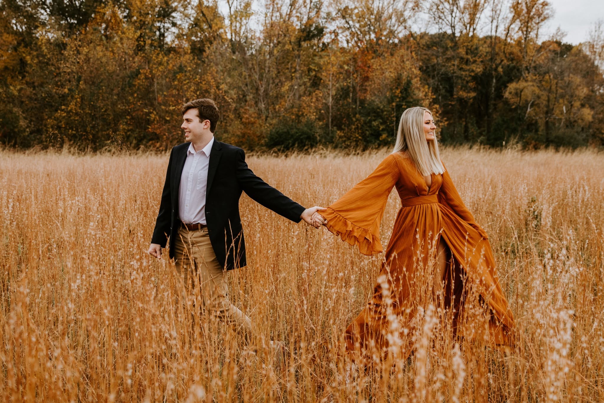 A man and woman holding hands in a field with trees in the background, woman wearing an orange dress.