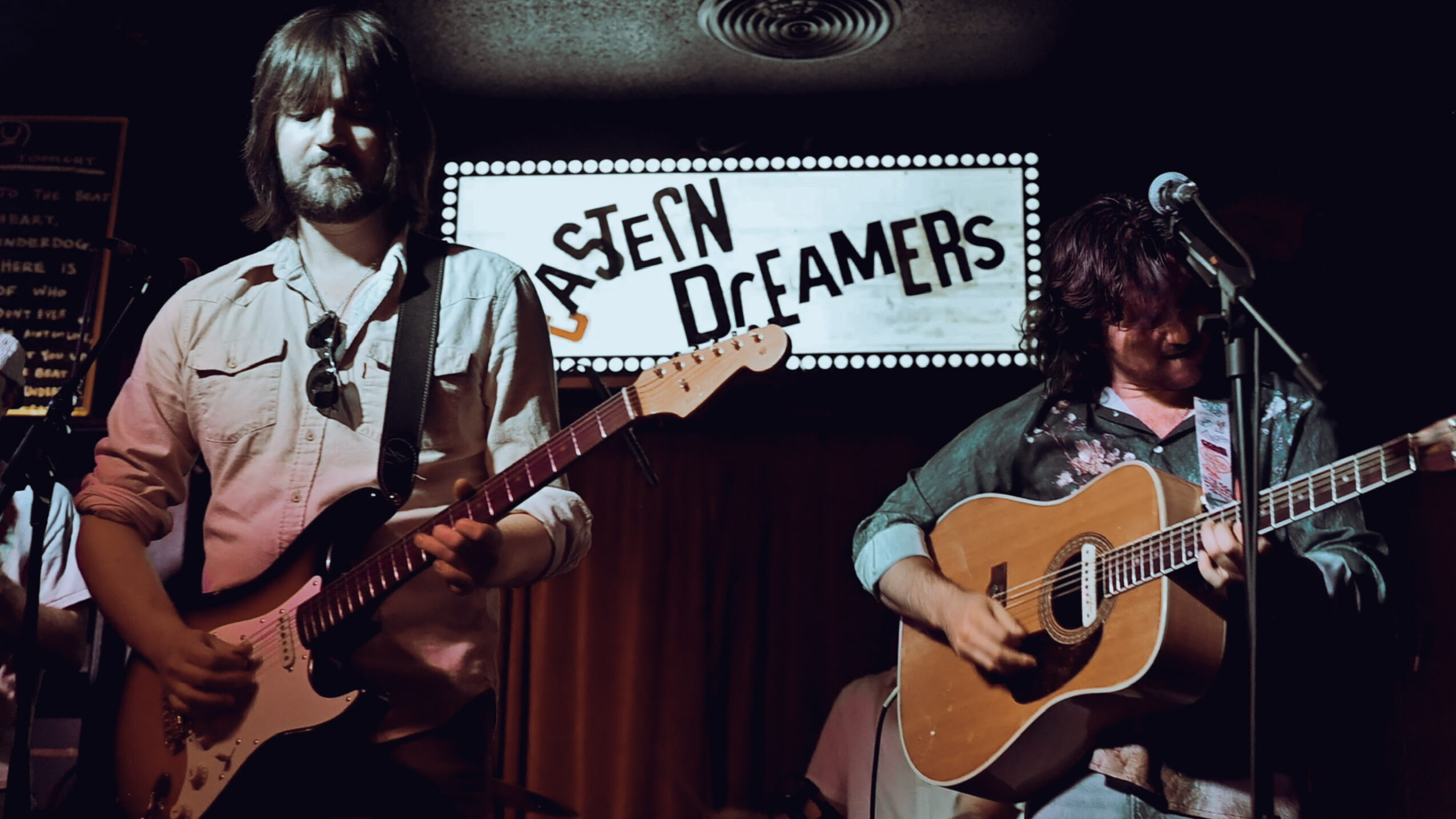 Two musicians perform on stage with guitars, a sign reads 'Asen Dreamers' behind them, dim lighting, curtain backdrop.