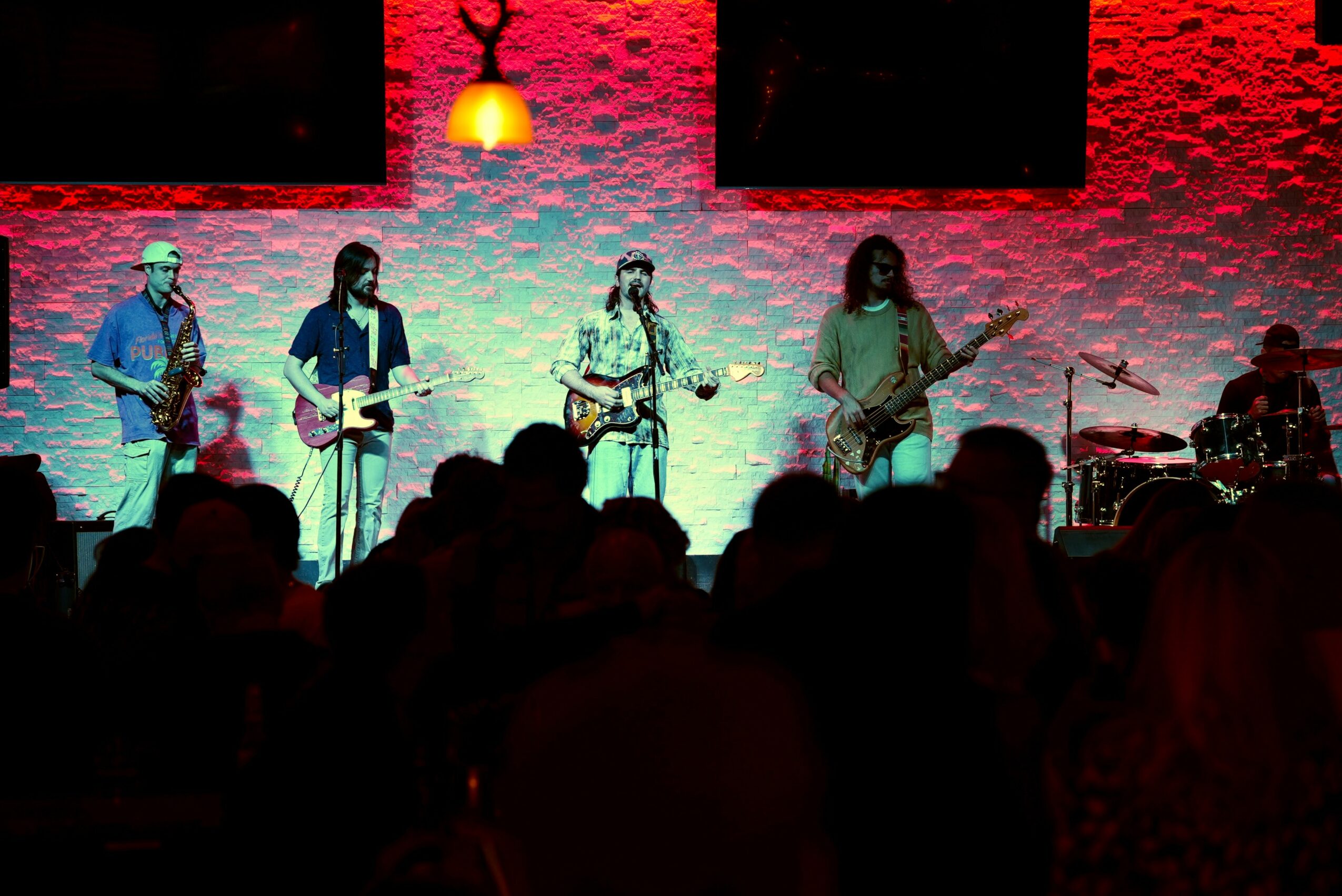 Band performs on stage with colorful lighting, audience silhouettes in foreground, brick wall background, and a hanging lamp.