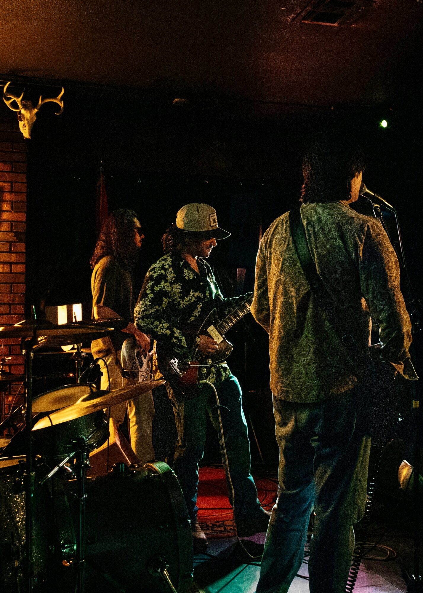 Three musicians perform on stage in a dimly lit venue, with a brick wall and deer antlers in the background.