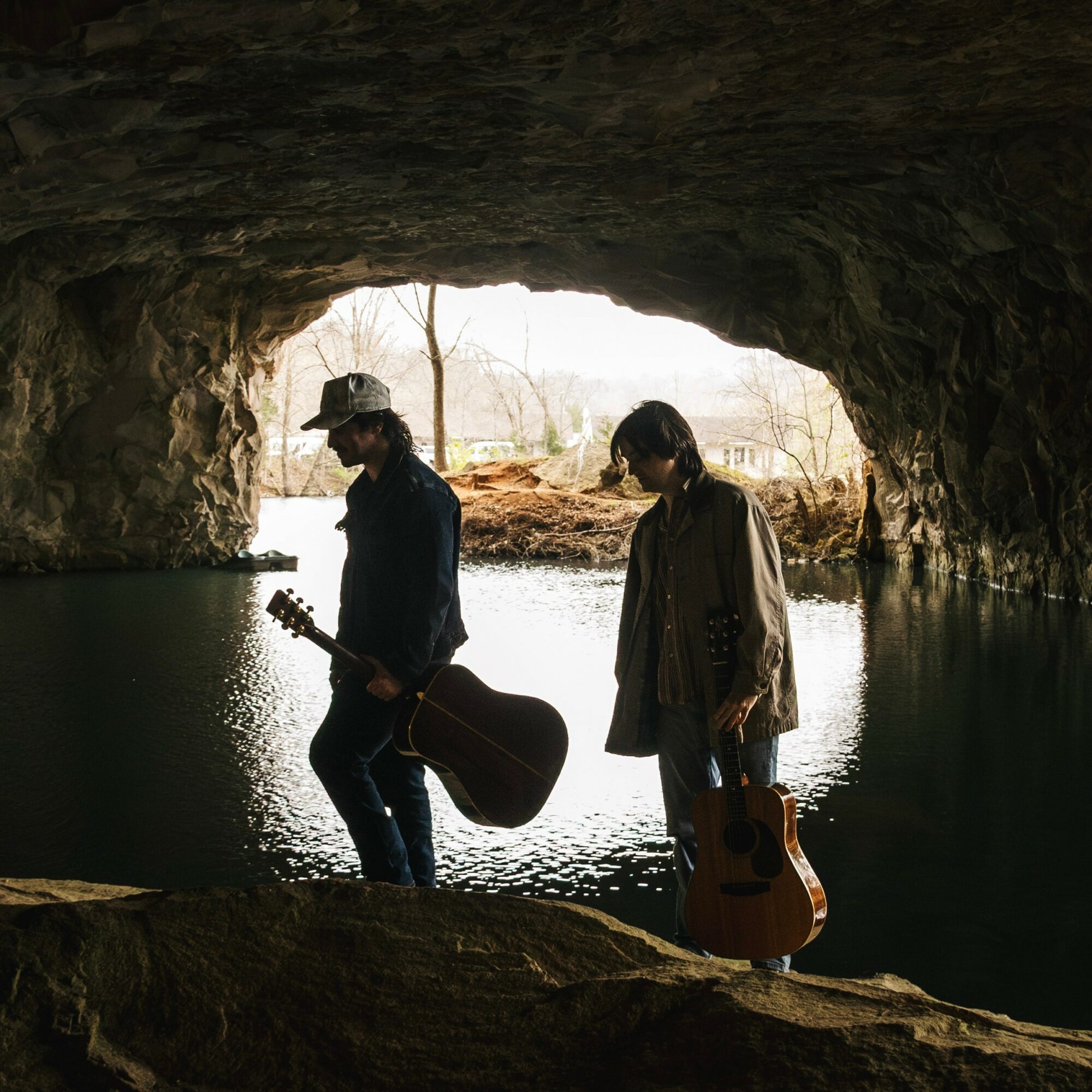 Two people with guitars standing in a cave opening overlooking a body of water, backlit by daylight.