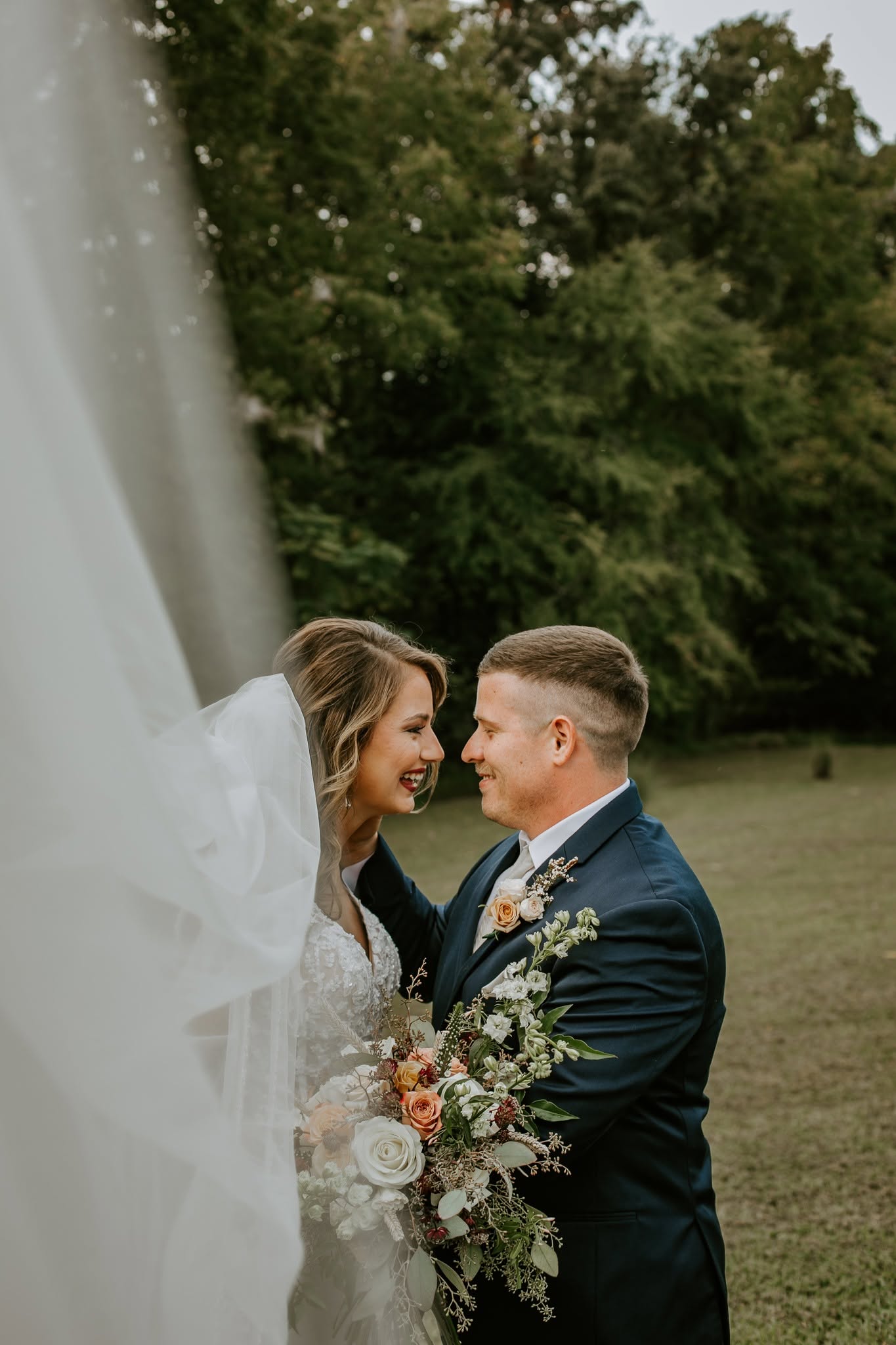 Bride and groom smiling at each other outdoors, bride holding a bouquet, trees in background.