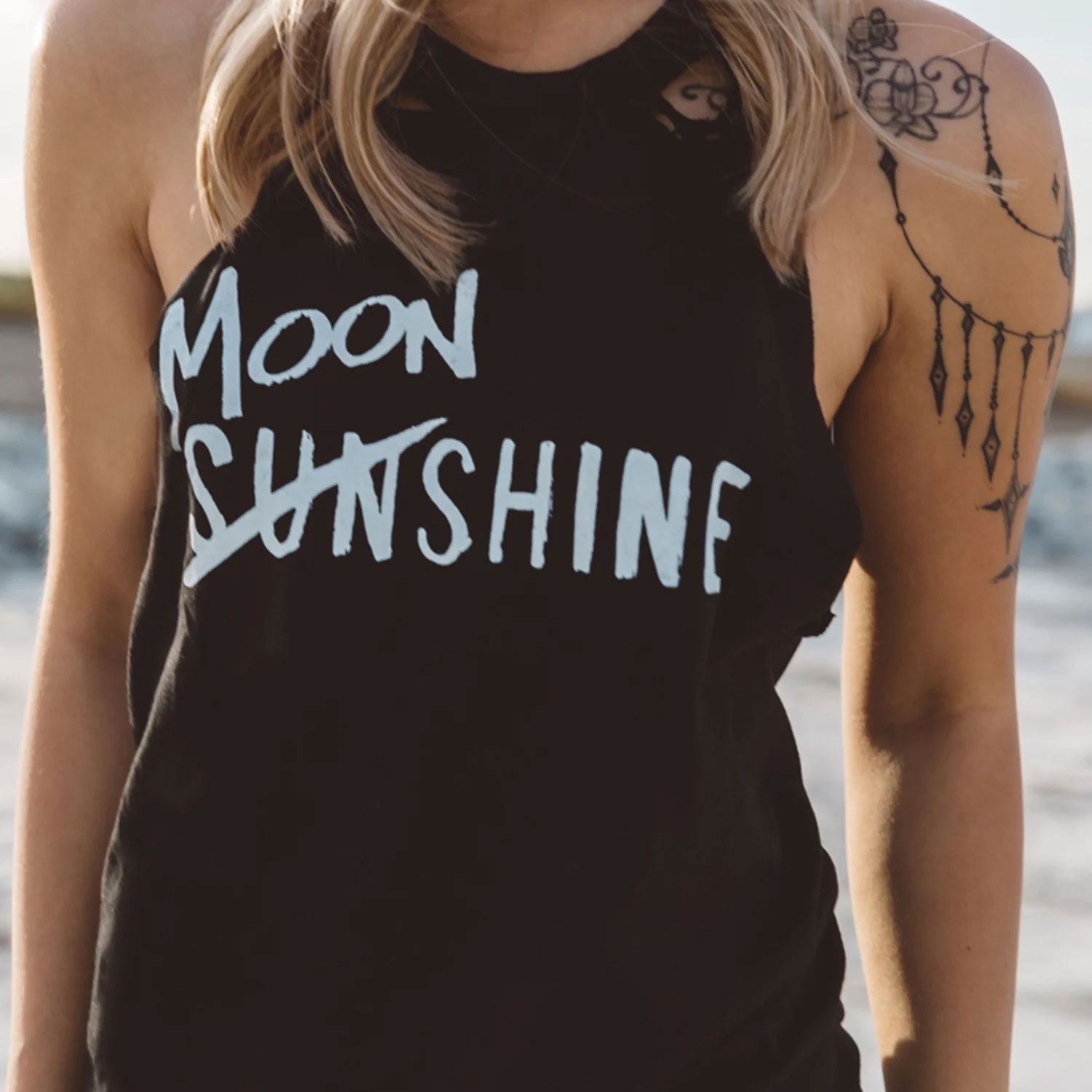 Person wearing a black sleeveless top with white text, showing tattoos on their shoulder and arm, outdoors.