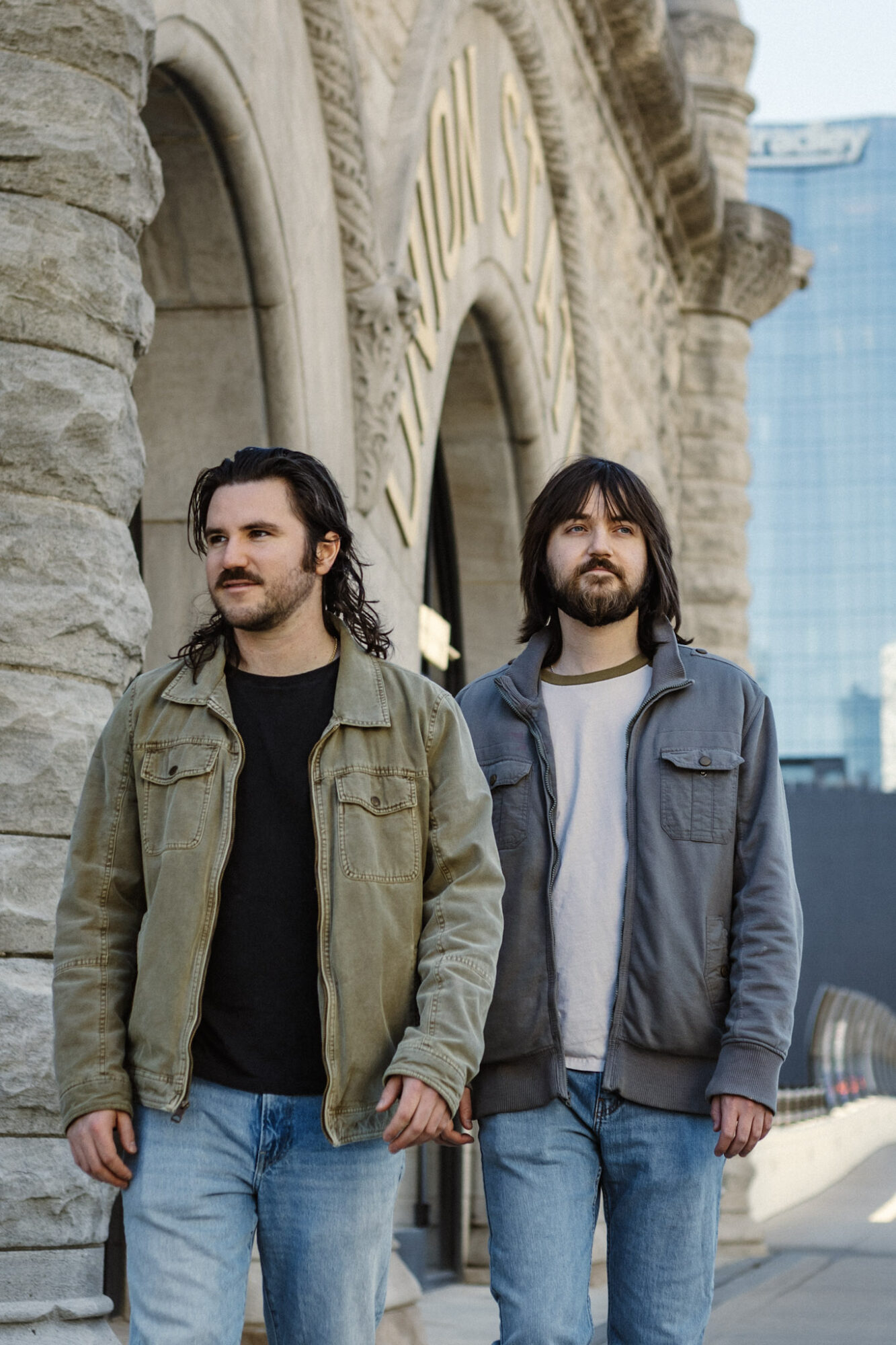 Two men with long hair and beards stand outside a stone building with arches, cityscape in background.