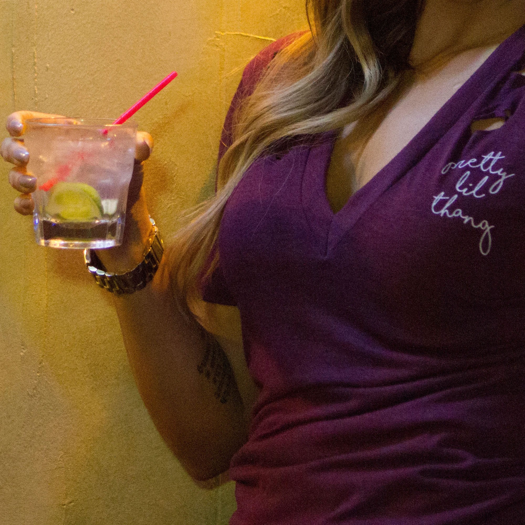 Person holding a drink with lime and straw, wearing a purple top with white text, long hair over shoulder.