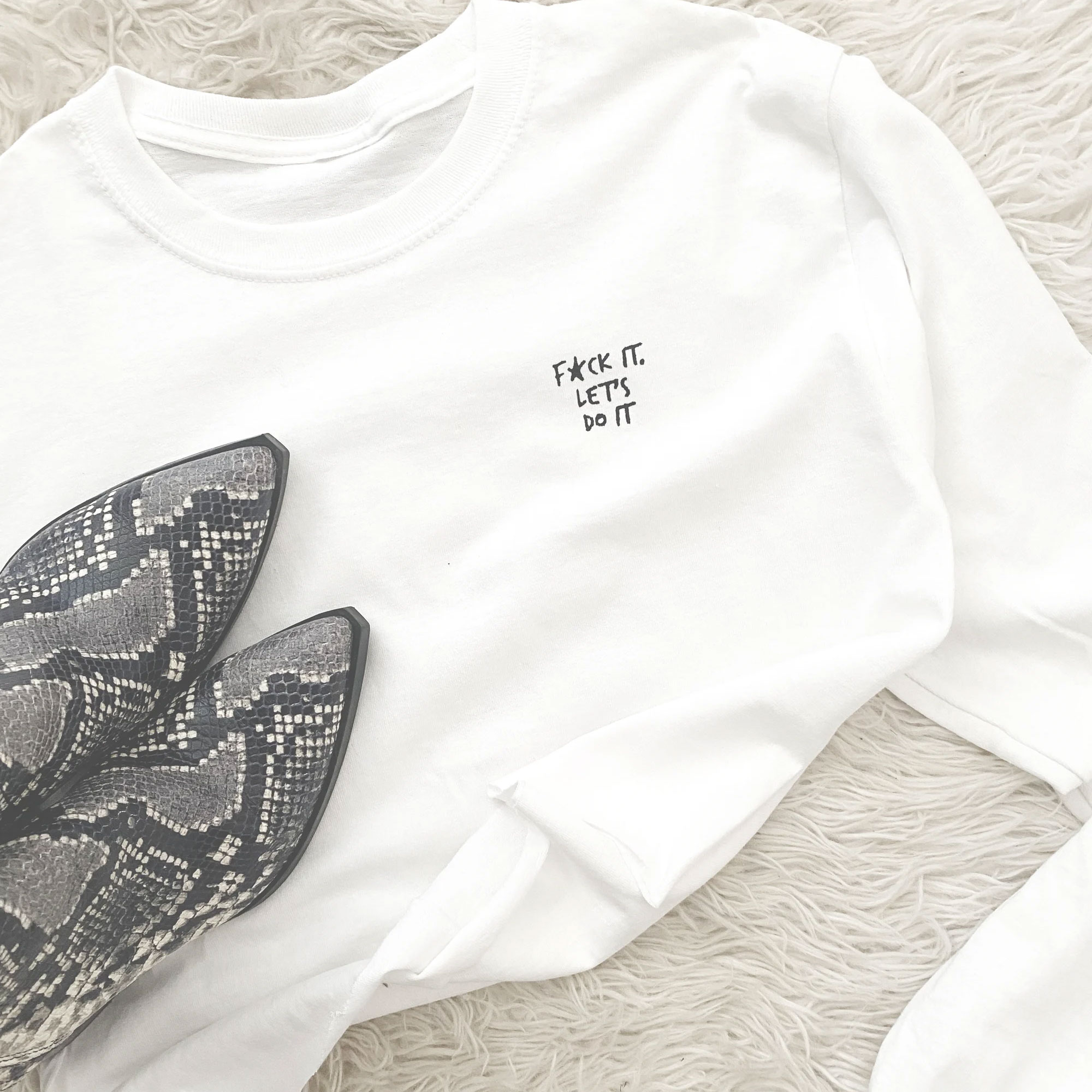White sweatshirt with small text reading 'F*** it, let's do it' and a pair of snakeskin-patterned shoes on a soft surface.