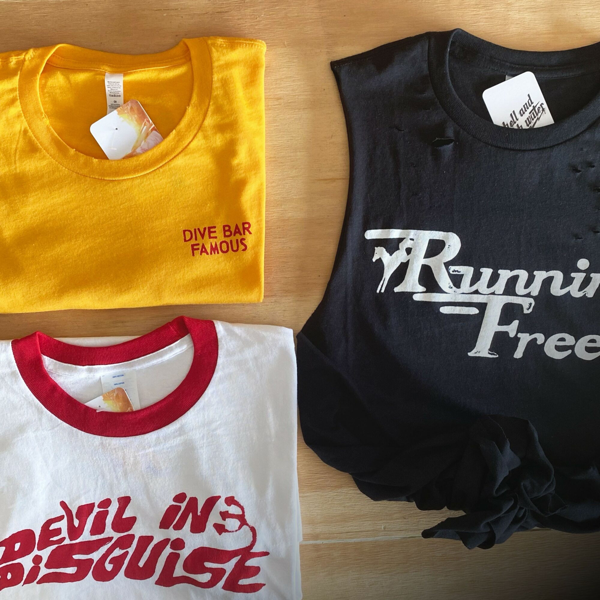 Three folded T-shirts on a wooden surface, yellow with red text, black with white text, and white with red text.