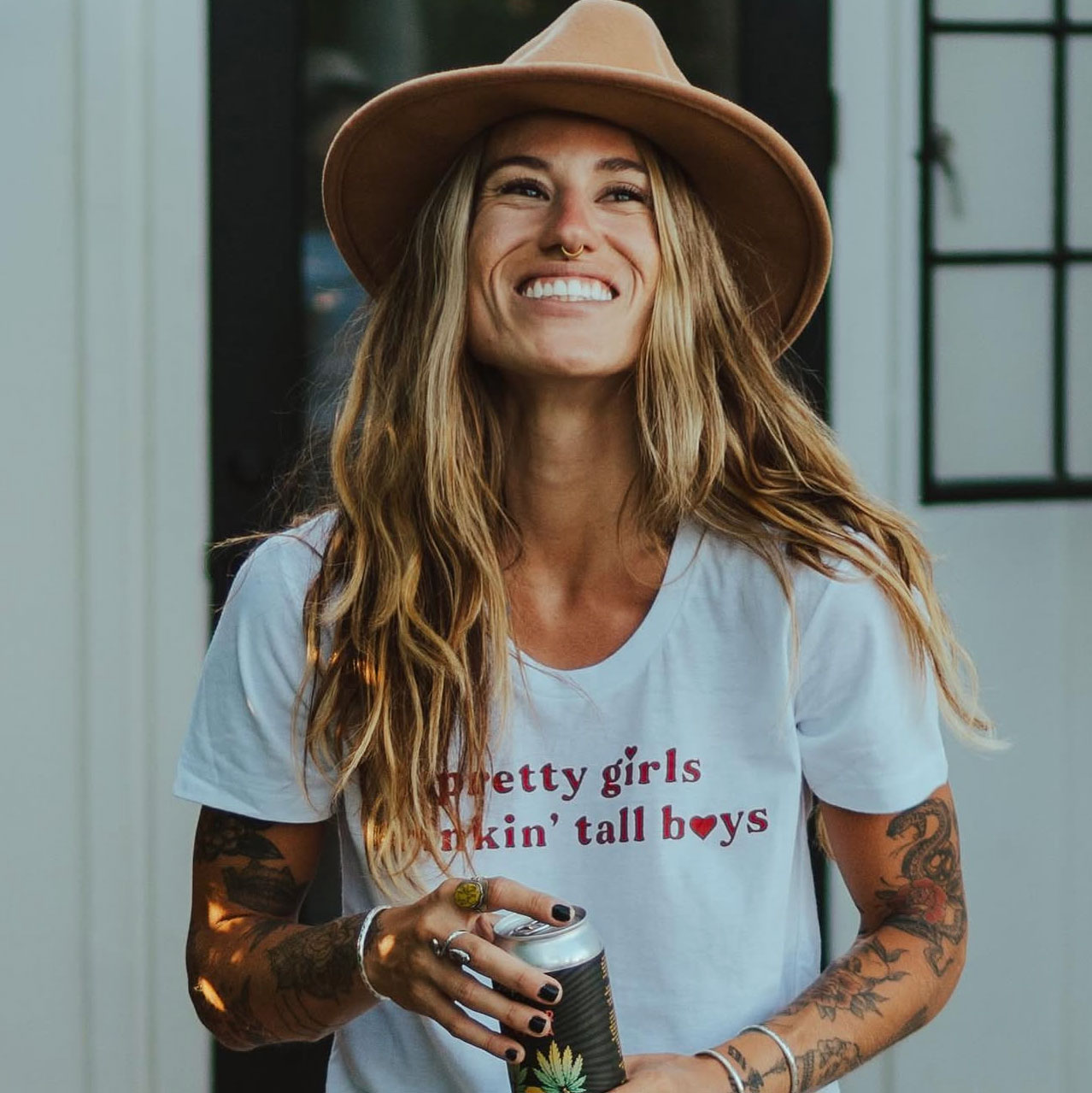 Smiling woman with long hair wearing a hat and a T-shirt, holding a camera, standing indoors near windows.
