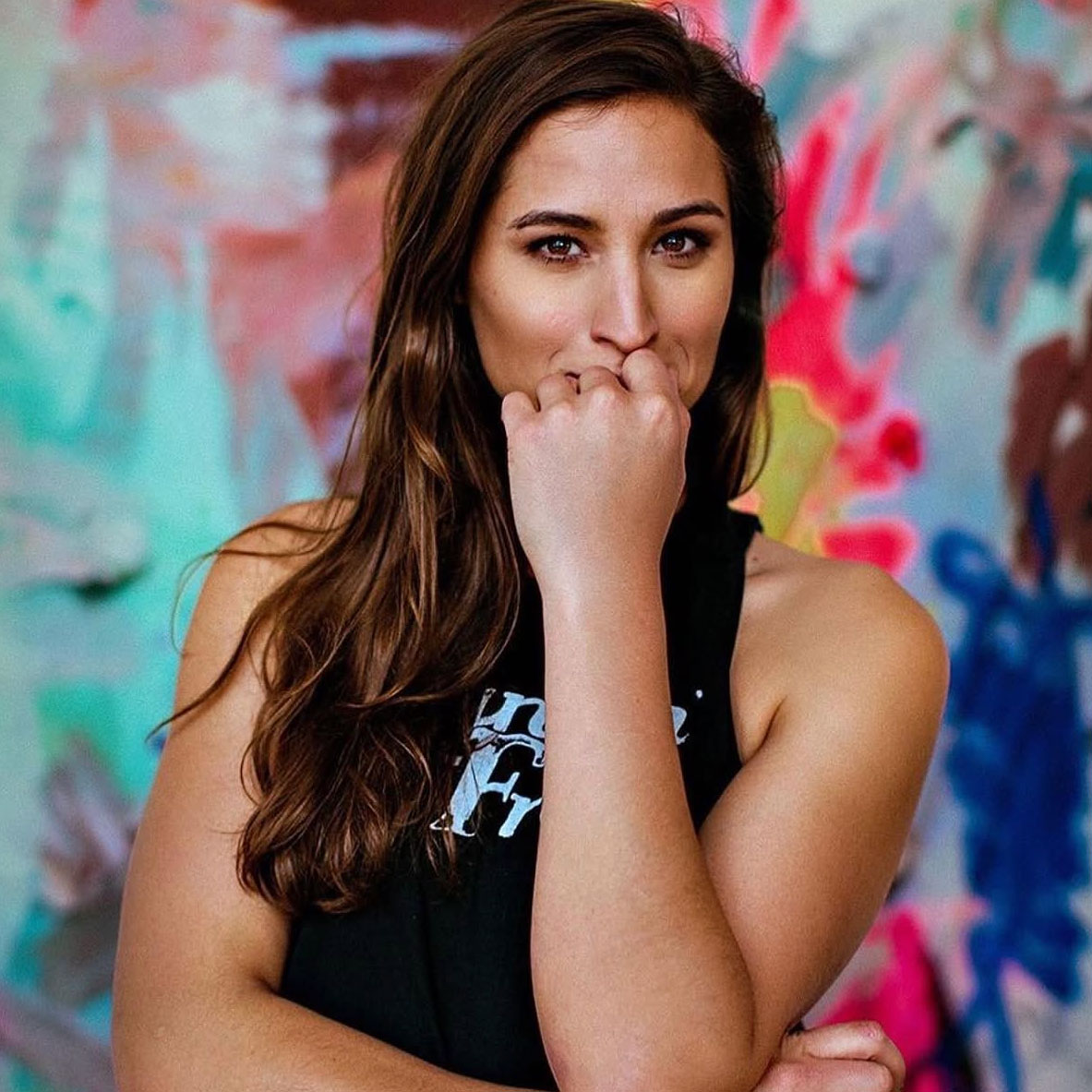 Young woman with long brown hair covering her mouth with her hand, standing in front of a colorful abstract background.