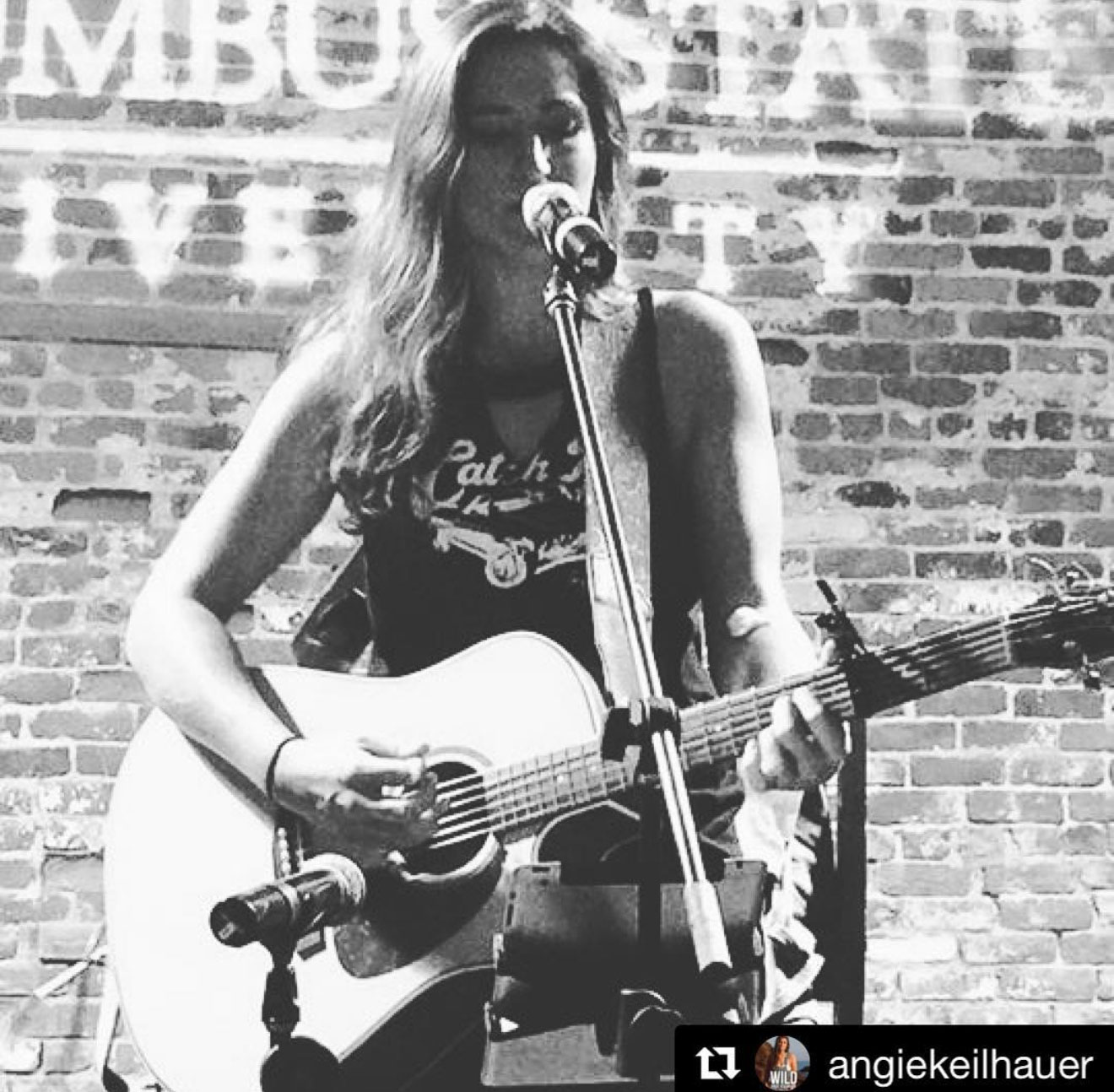 Woman playing guitar and singing into microphone on stage with brick wall background.