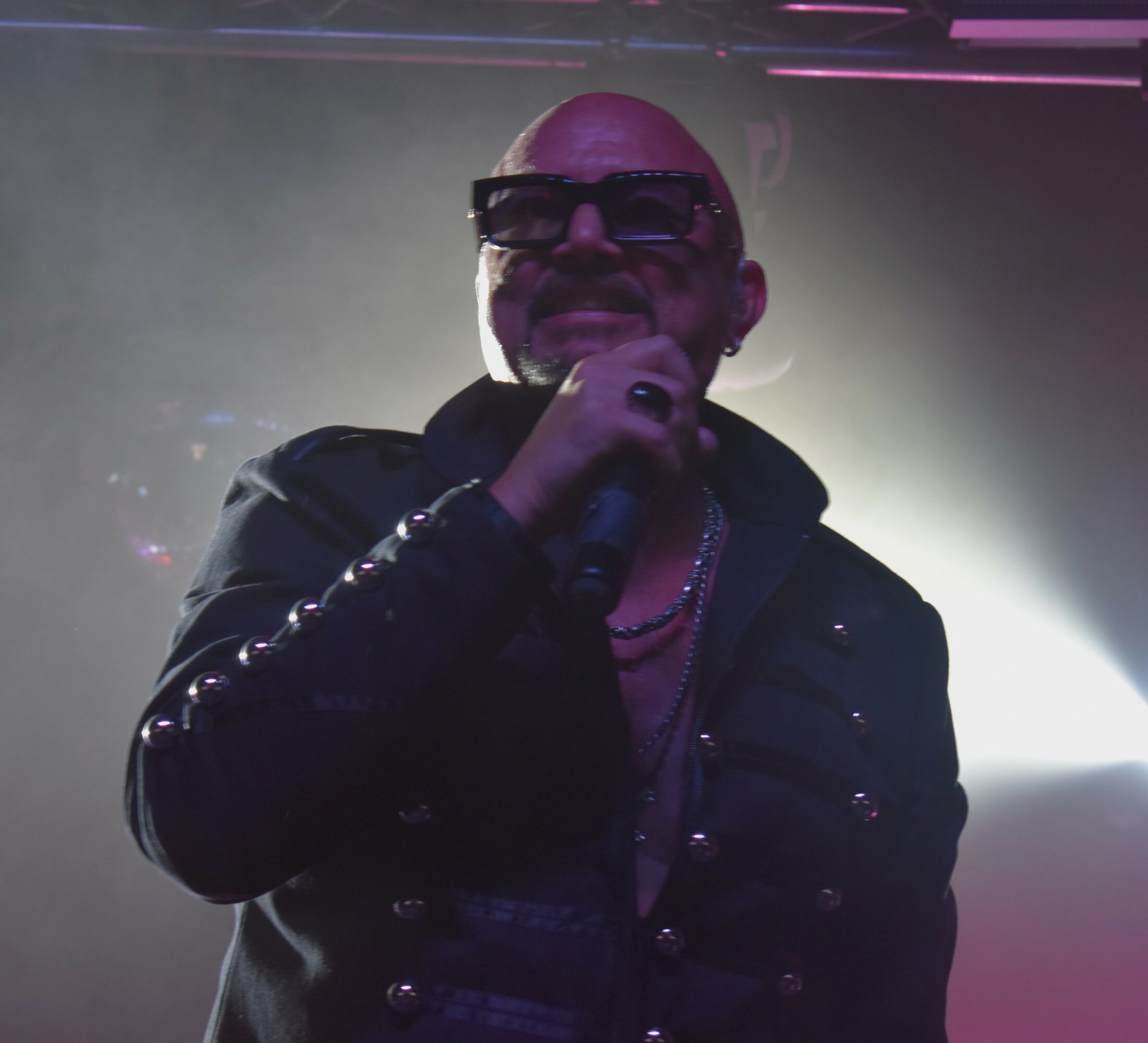 Bald person wearing sunglasses and a dark jacket, holding a microphone, with stage lighting behind.