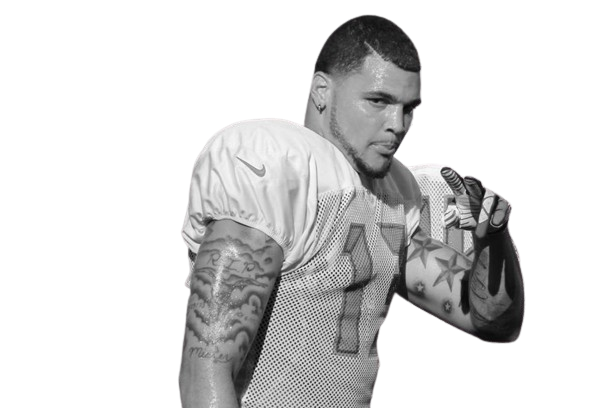 A football player in uniform with tattoos on his arm, holding a helmet, against a black background.