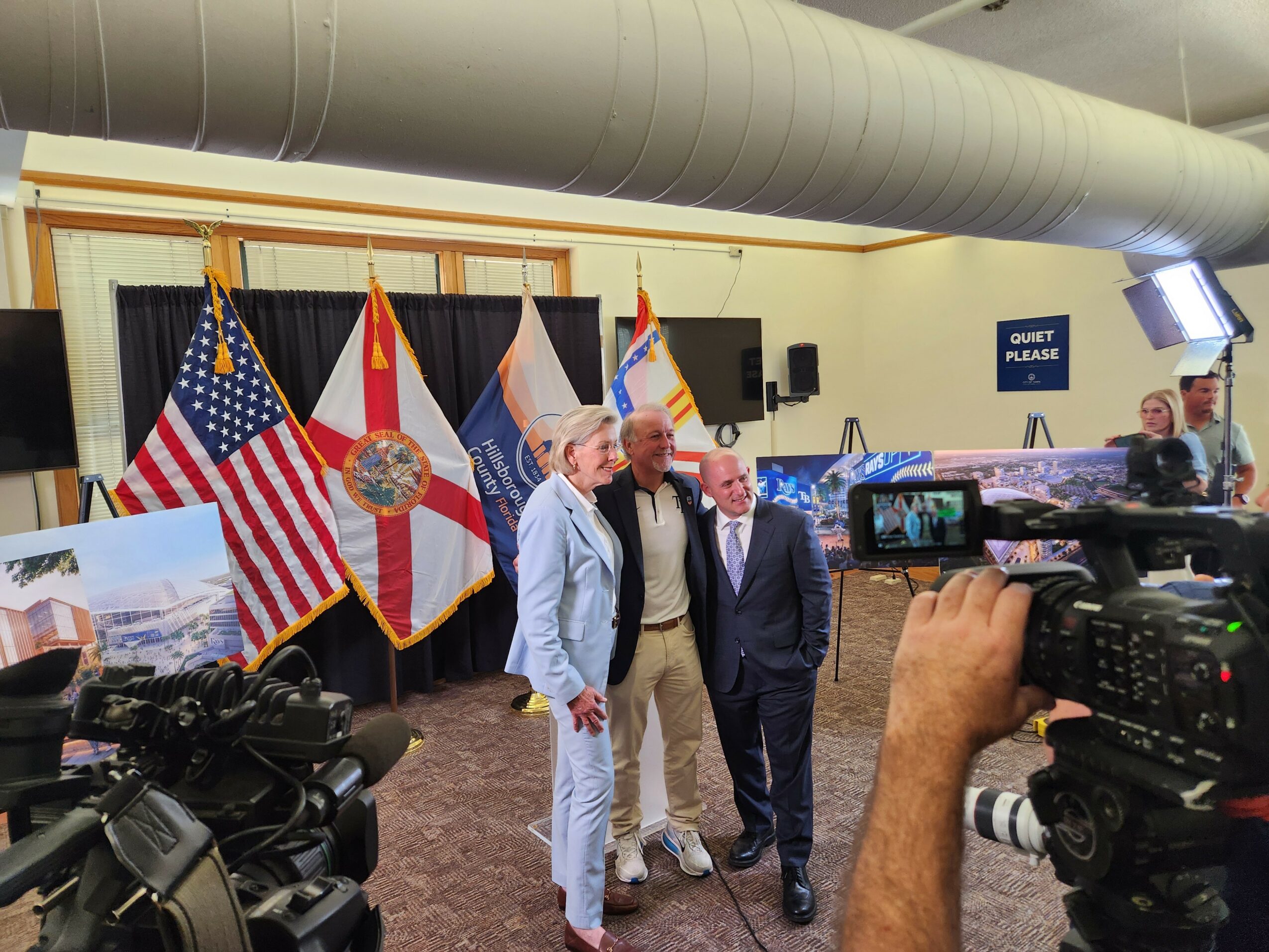 Three men stand together in front of flags, with cameras recording the scene, in an indoor event space.