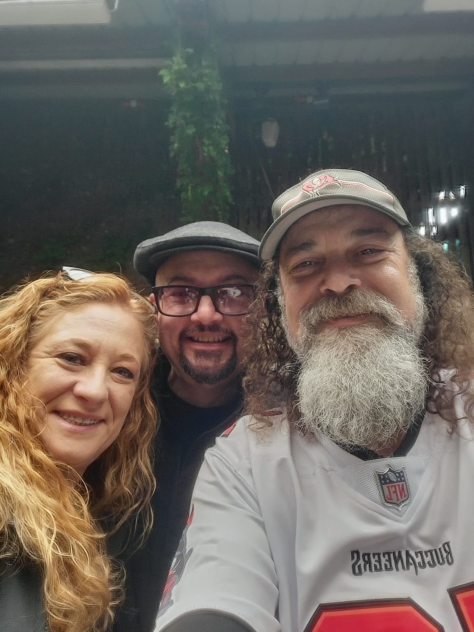 Three people smiling closely for a selfie outdoors, with a dark background and some greenery visible.