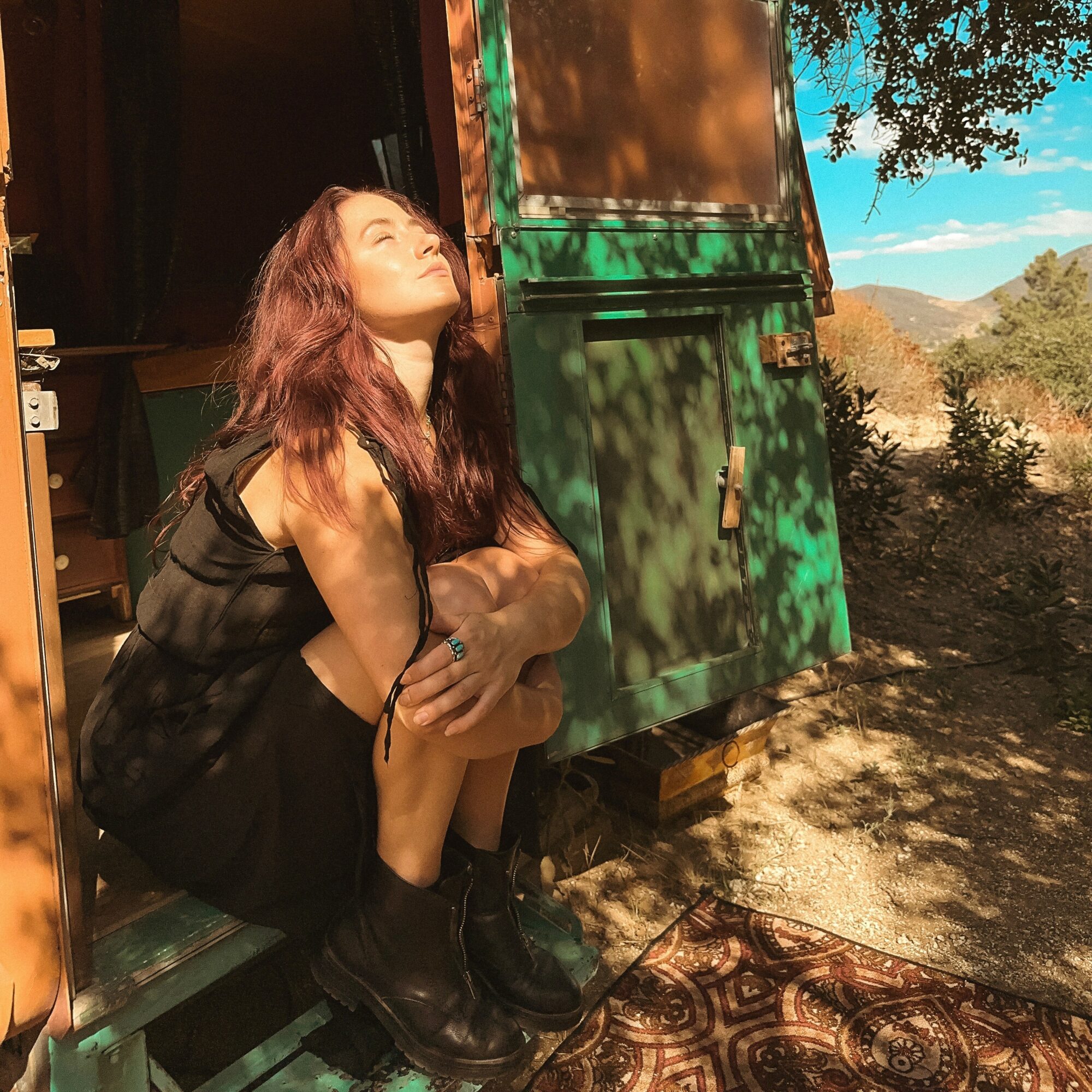 Young woman with long hair sitting outside near a green structure, sunlight on her face, landscape in background.