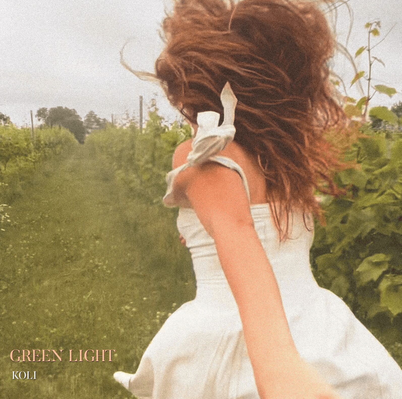 Person with curly hair wearing a white dress outdoors, surrounded by greenery, facing away from the camera.