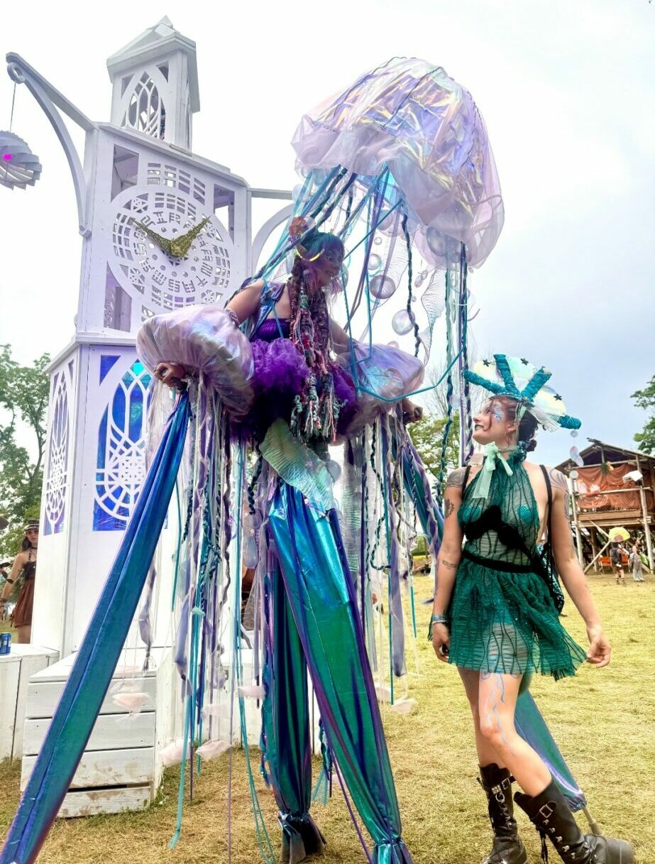 Person in a green dress and headpiece standing near a large, colorful, decorated structure outdoors.