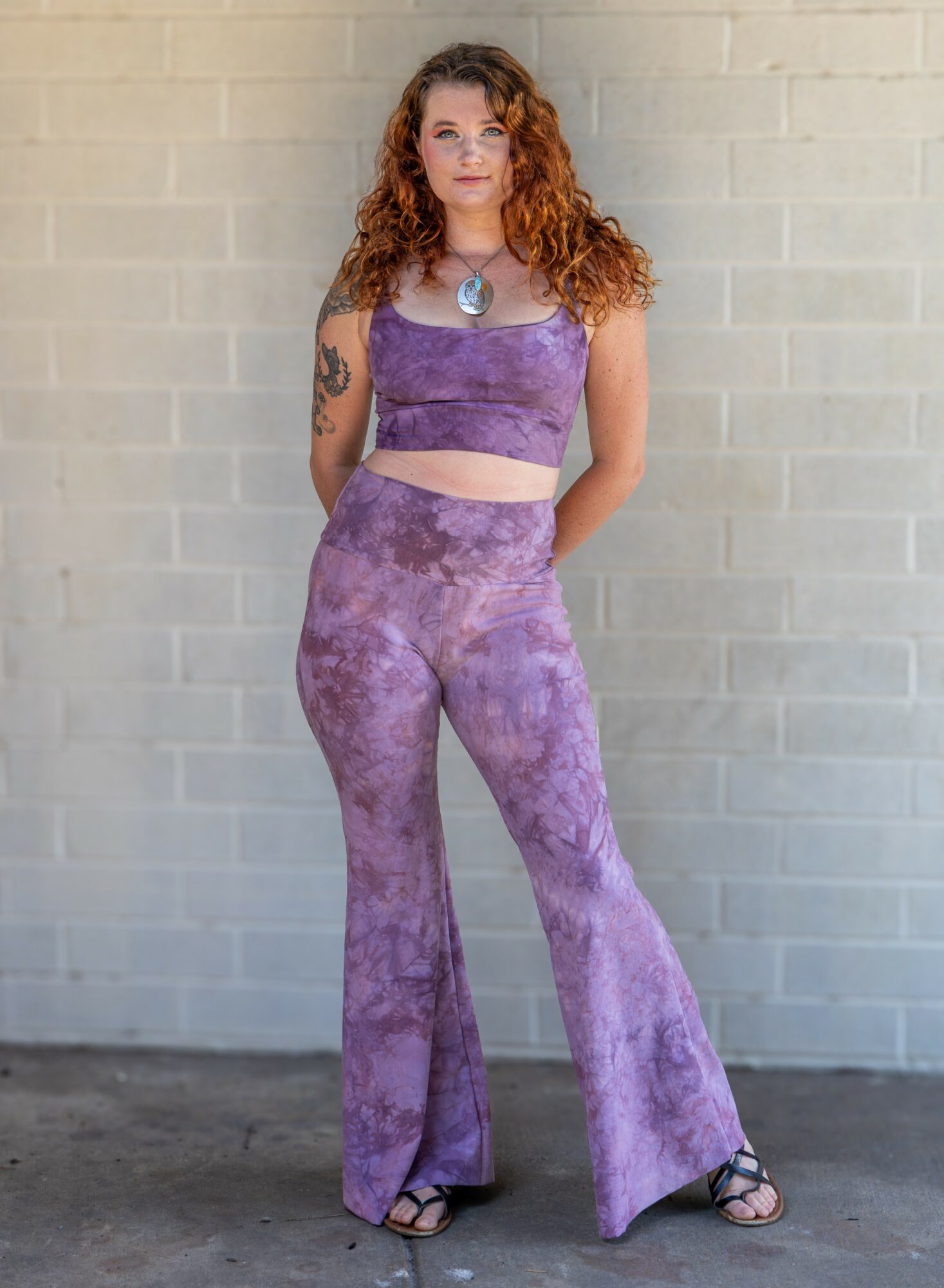 Woman with curly hair in purple tie-dye outfit stands against a light brick wall.