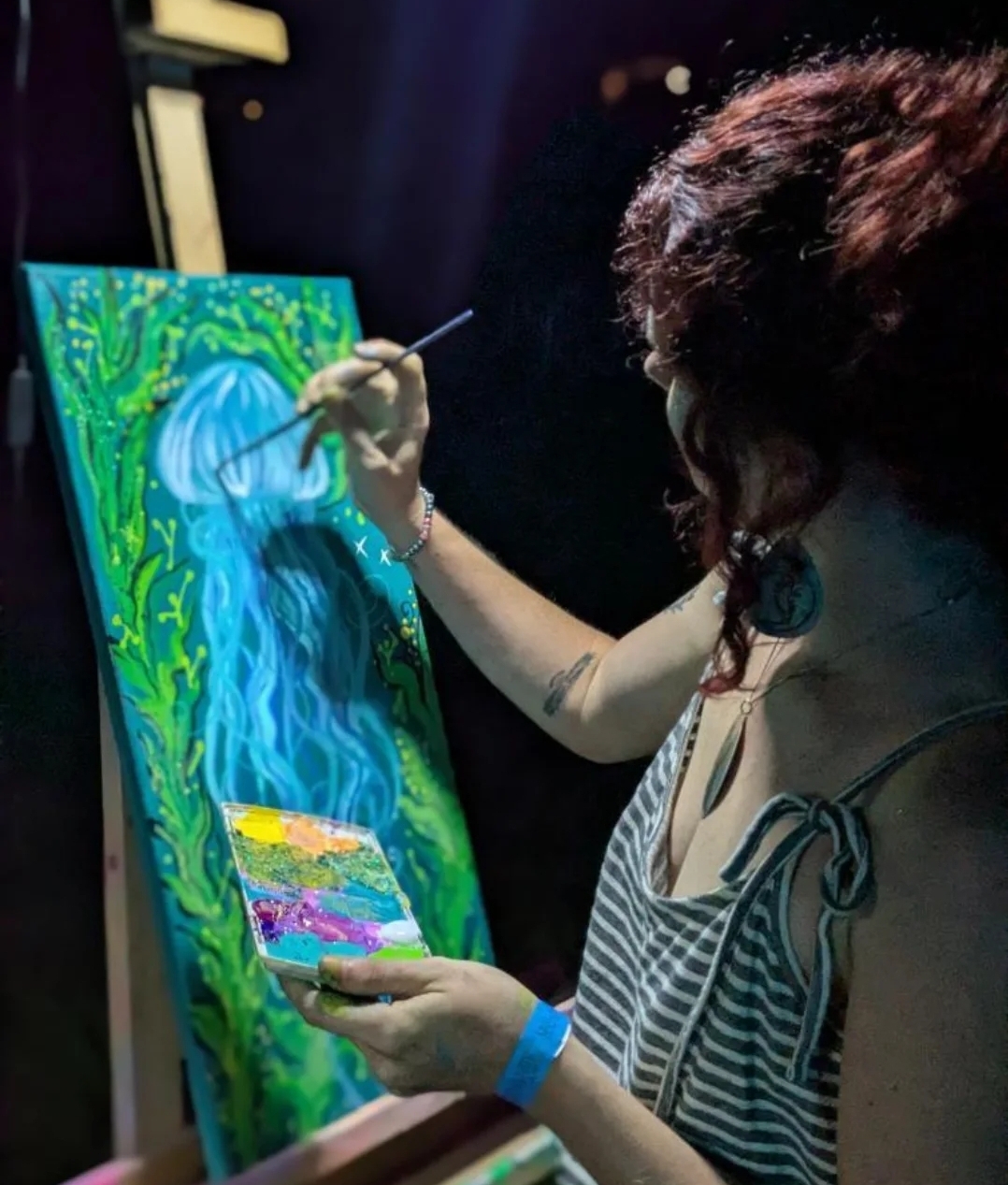 Person painting a waterfall on a tall canvas, holding a palette, with dark background and striped top.