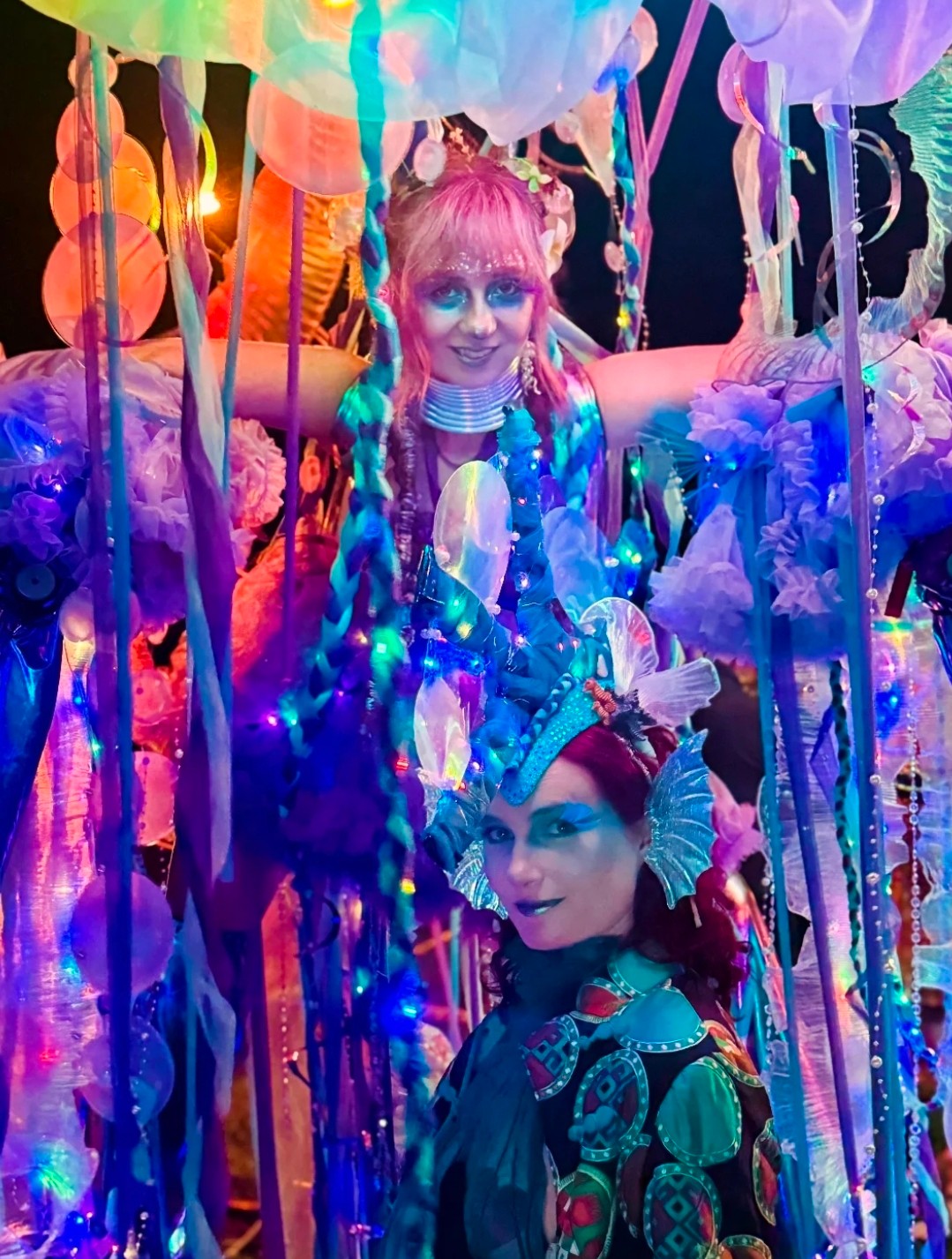 Two women in colorful costumes surrounded by vibrant decorations and hanging streamers, smiling and posing for the camera.
