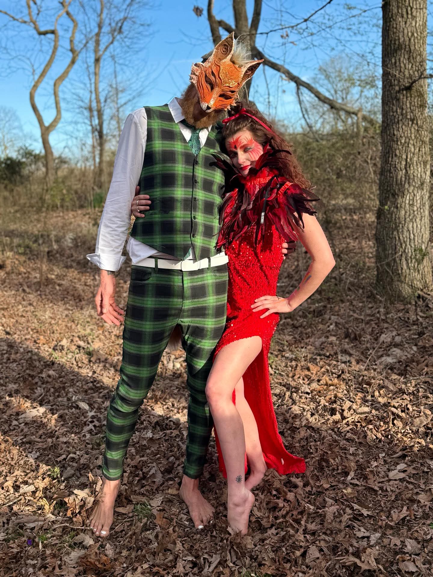 Two people in costumes stand outdoors on fallen leaves, one wearing a fox mask and the other in a red dress.